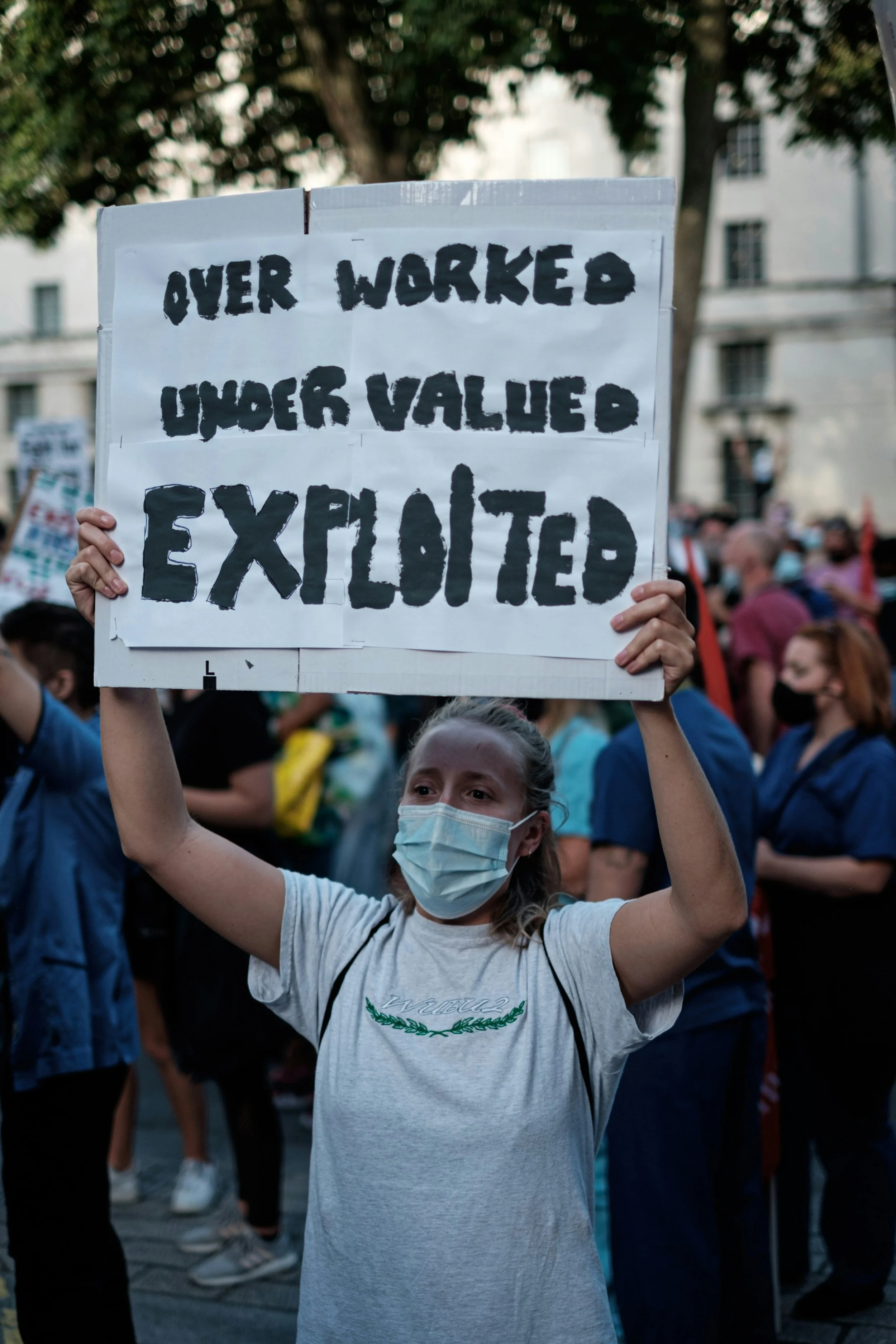 Woman in a white shirt and wearing a mask. She is holding a sign above her head that has three words: Overworked. Undervalued. Exploited.