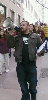 Chris Stout, union consultant & labor negotiator, walks along a picket line holding a megaphone to his mouth, and holding a clipboard in his other arm. c. 2009.