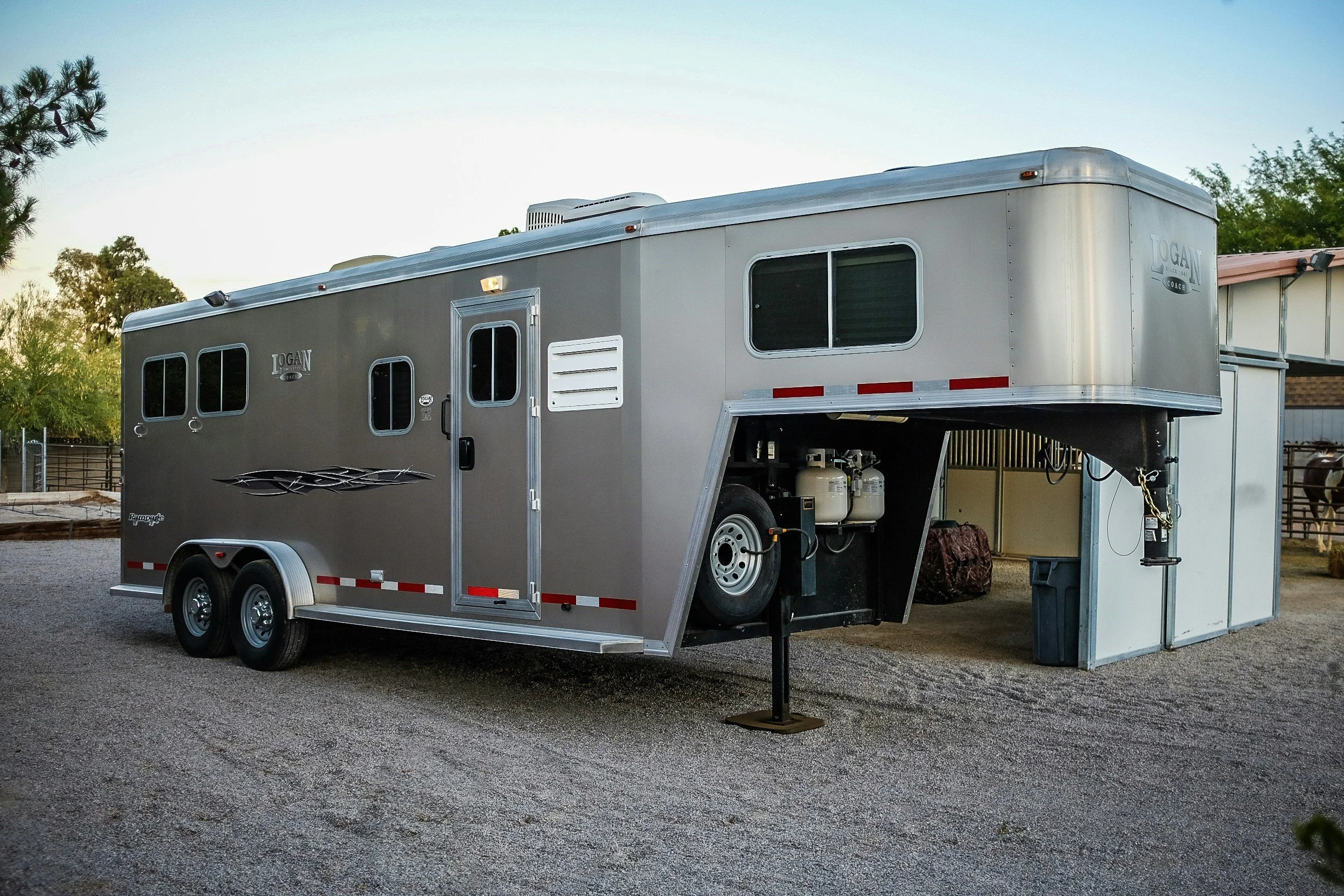 Silver horse trailer parked outdoors on gravel, with trees and a small building in the background.