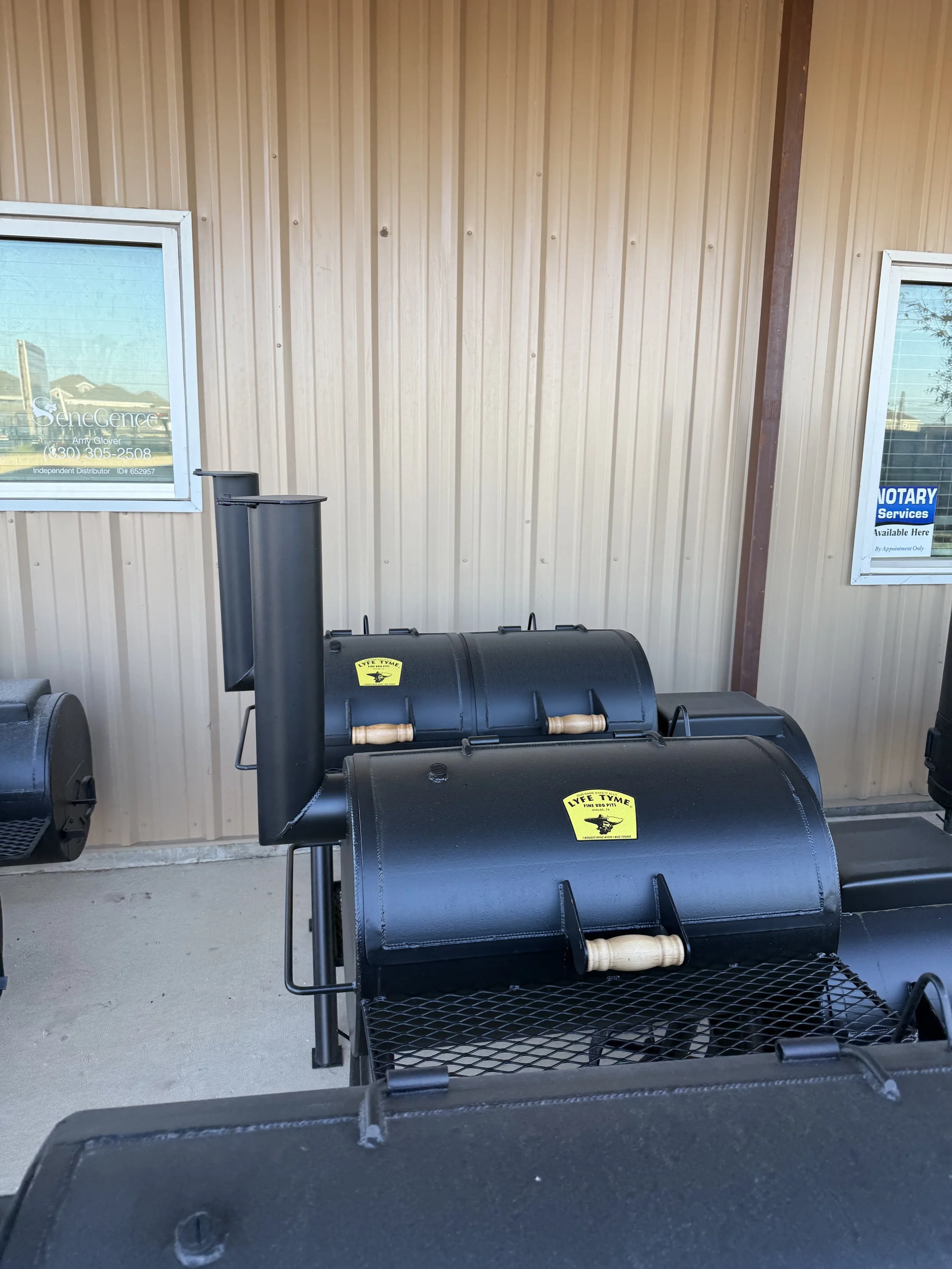 Two black barrel smokers with wooden handles and yellow stickers on a patio, against a beige metal building with two windows.