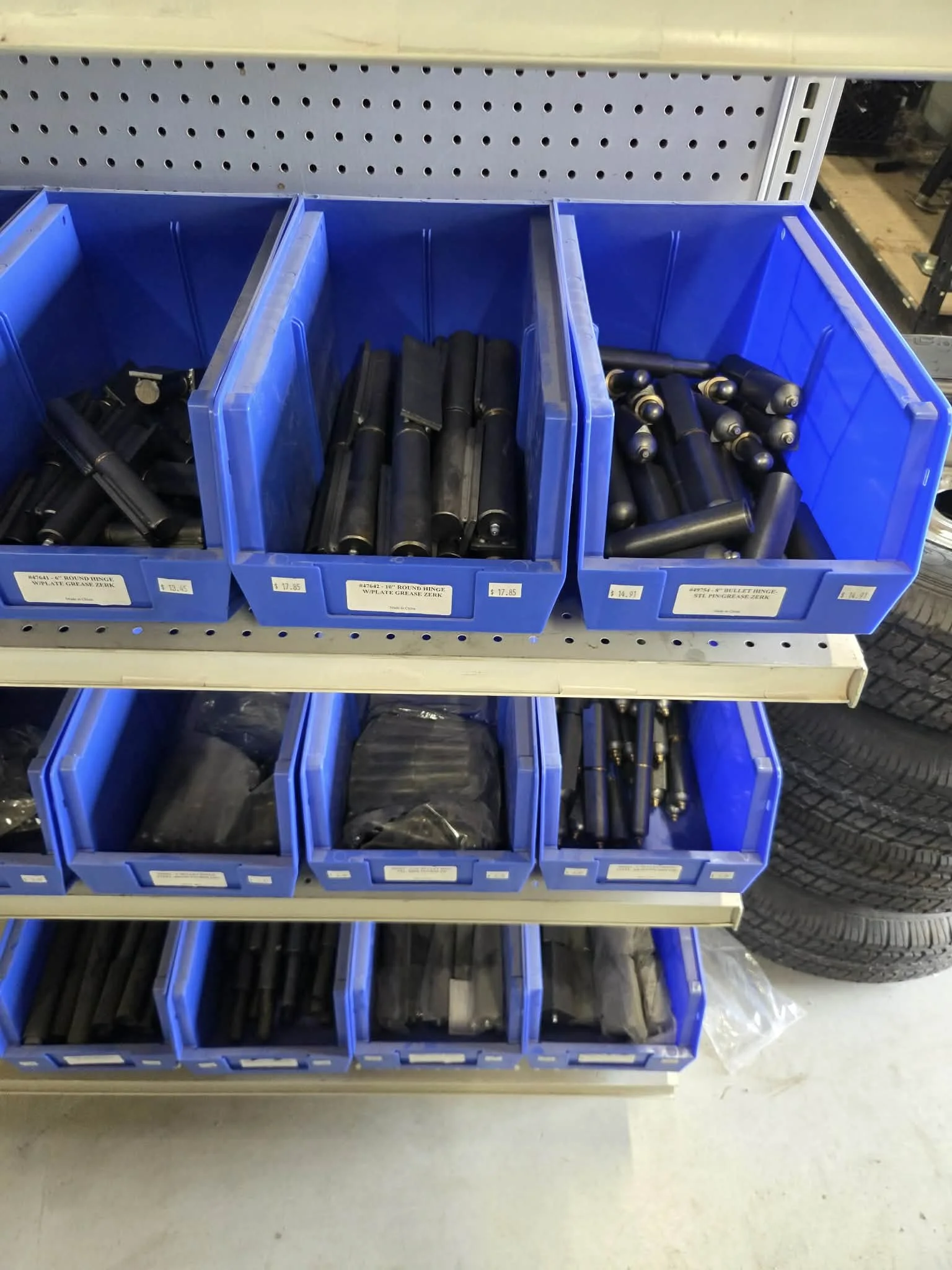Blue plastic bins on a metal shelving unit, containing black metal hardware parts, possibly for machinery or equipment. Labels with prices are attached to the bins.