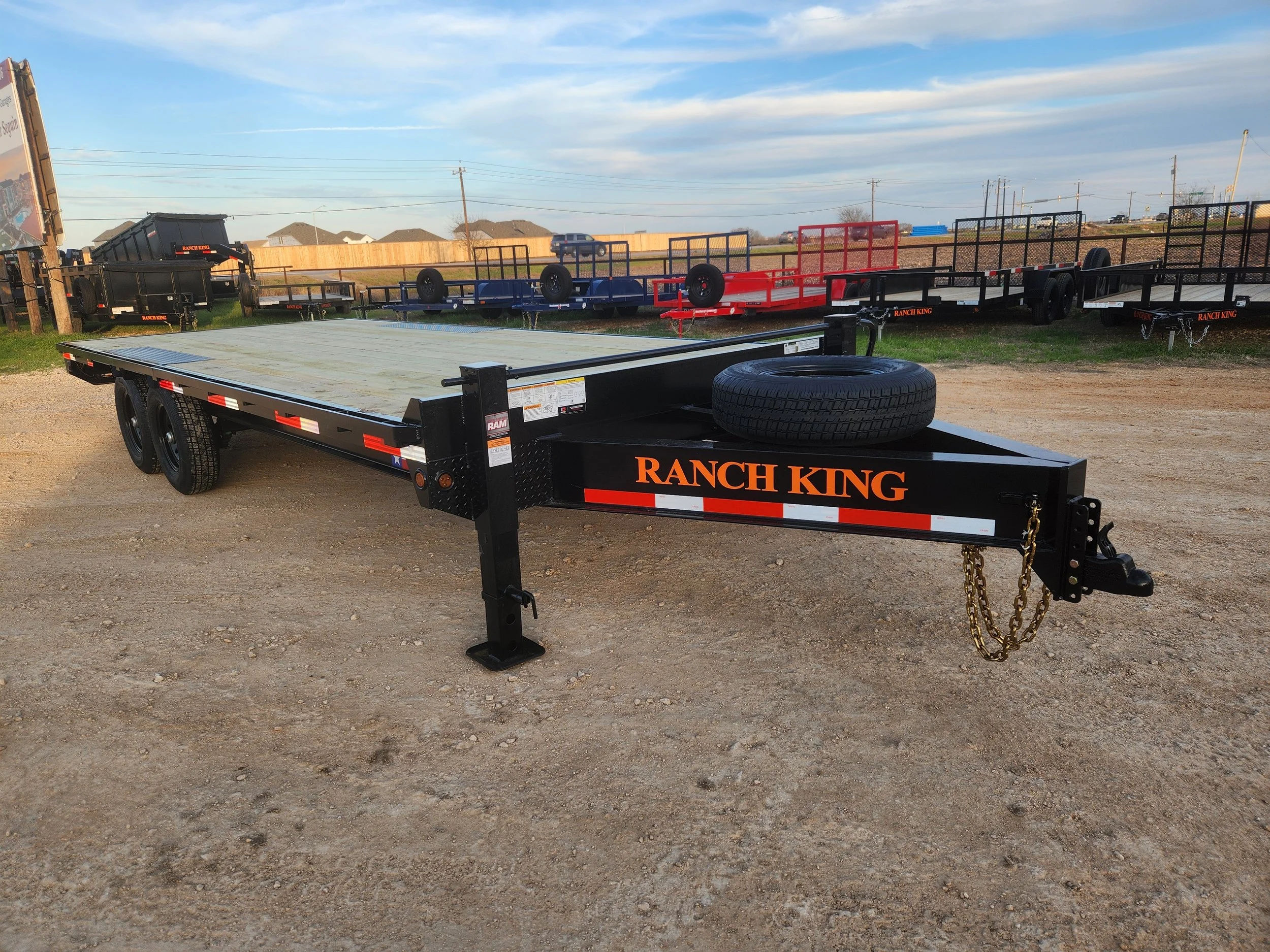 A black ranch king flatbed trailer with a spare tire mounted at the front, parked on a gravel lot, with other trailers in the background under a blue sky with clouds.