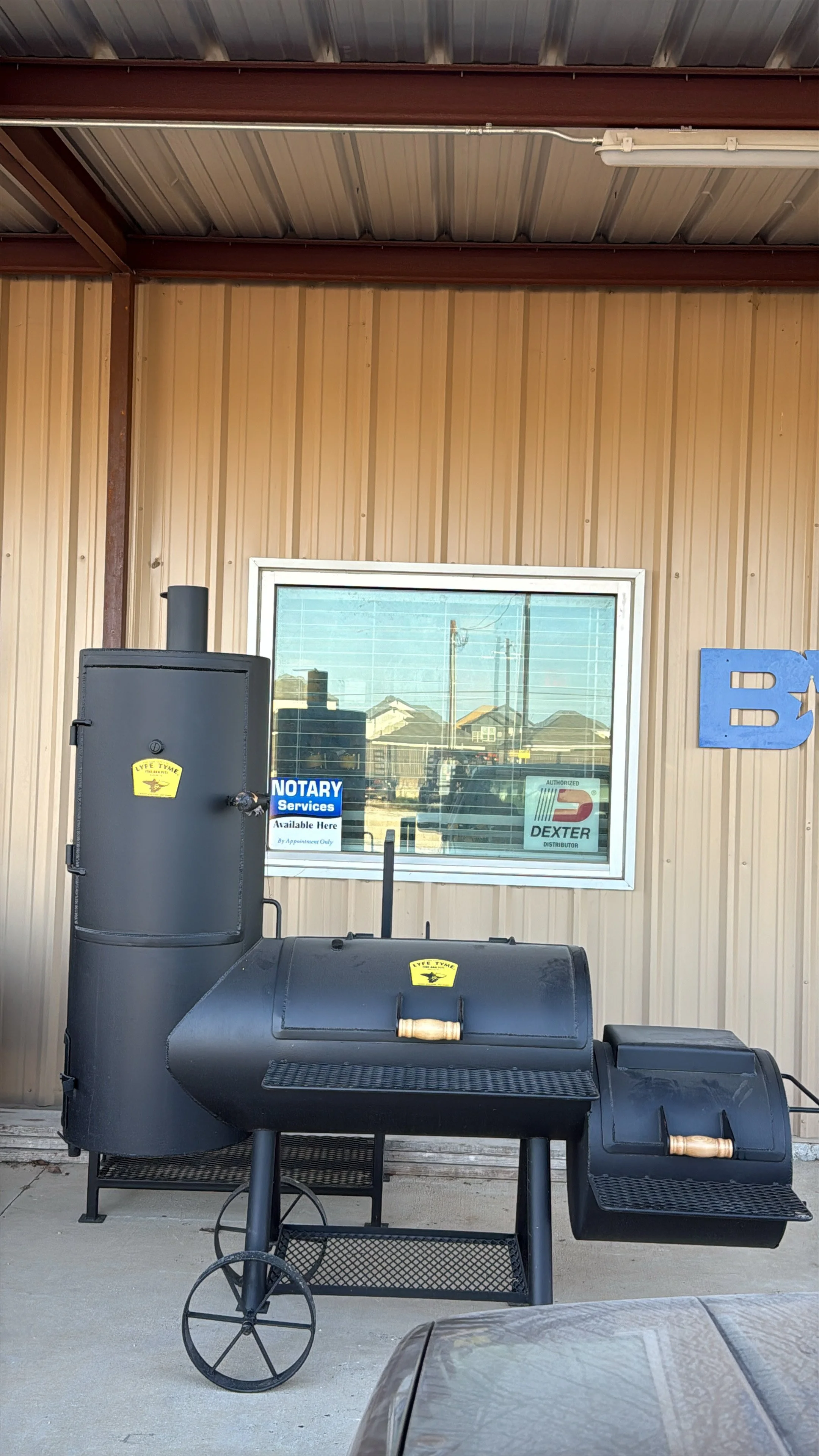 Black outdoor smoker grill with two chambers, positioned in front of a beige metal building, window with signs, and a partial view of a vehicle hood.