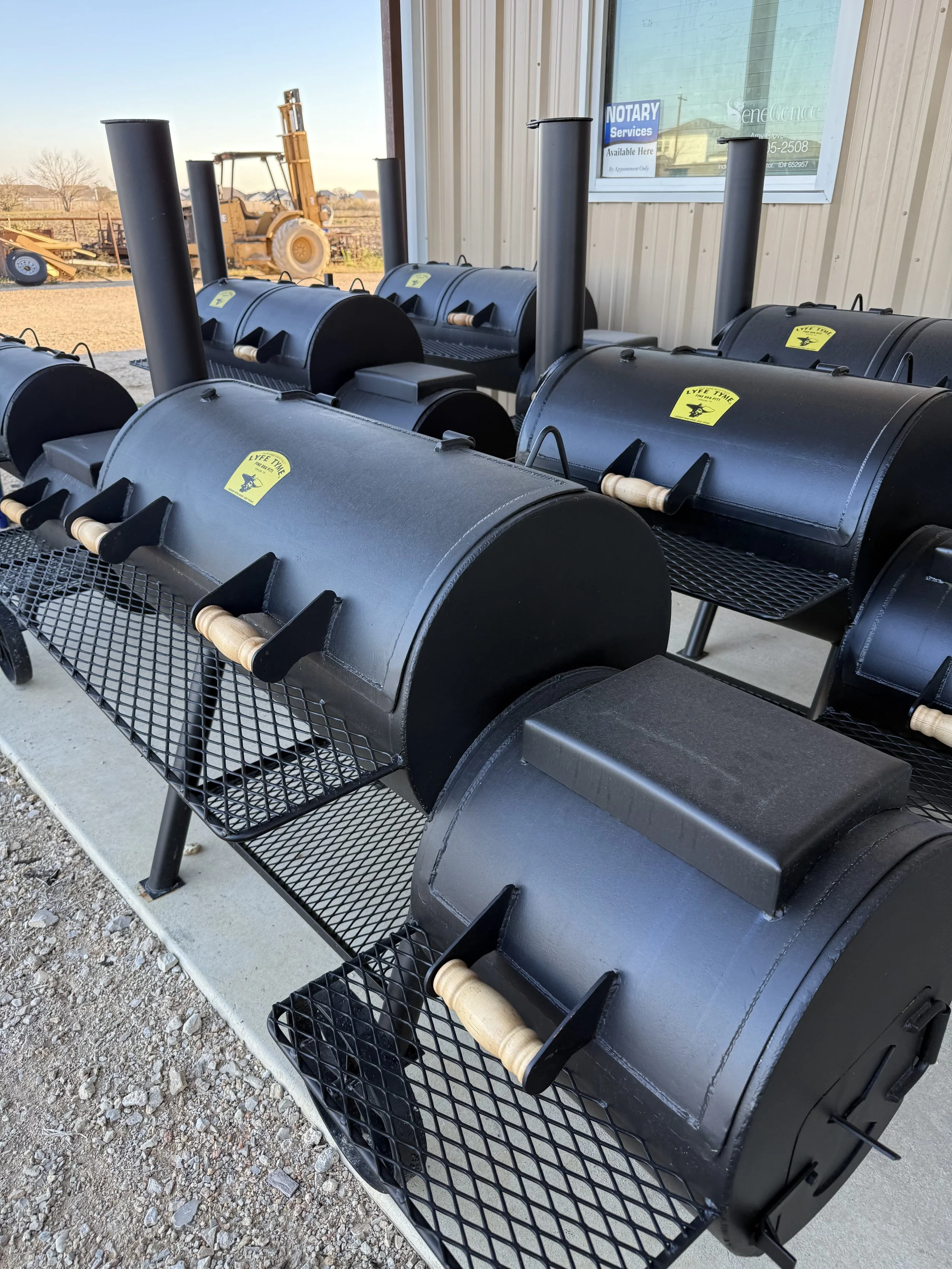 Several black barbecue smokers with wooden handles on a metal rack outside a building.