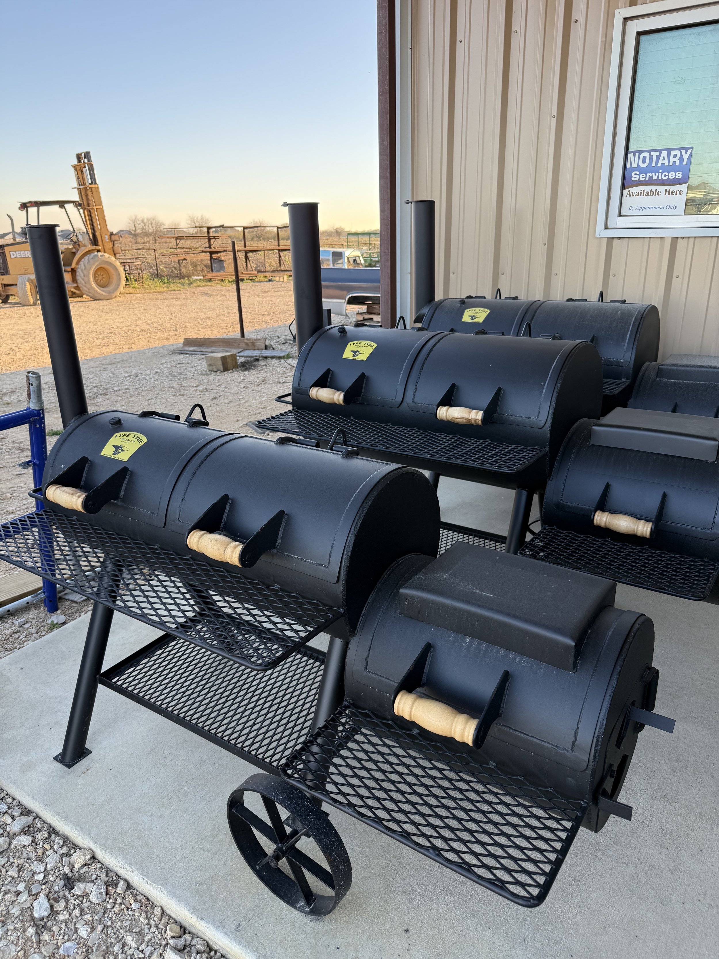 Three black barrel-style smokers with wooden handles on each, placed on a concrete surface outside a building with a beige siding wall and a window with a sign that reads 'NOTARY Service Available Here.' in a rural setting with a dirt lot and constru