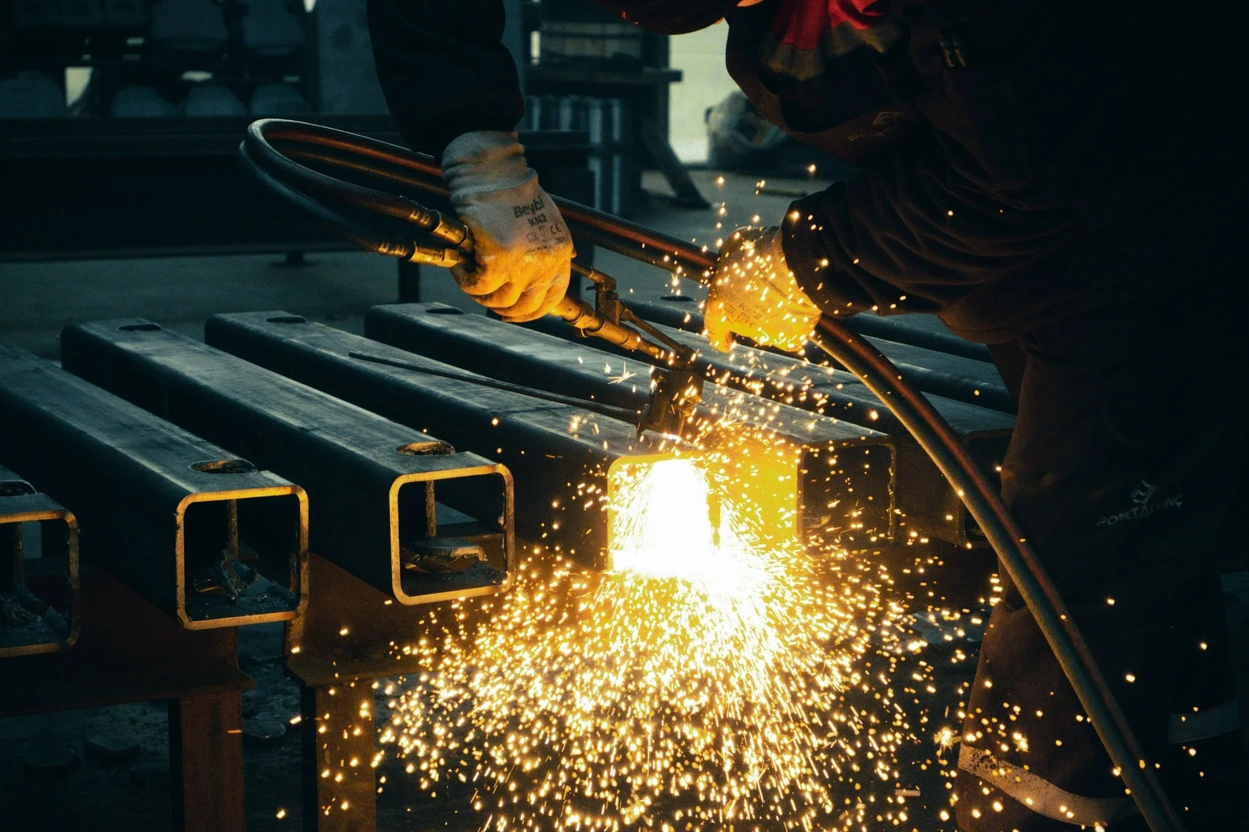 A worker welds metal pipes, sparks flying in an industrial workshop setting.