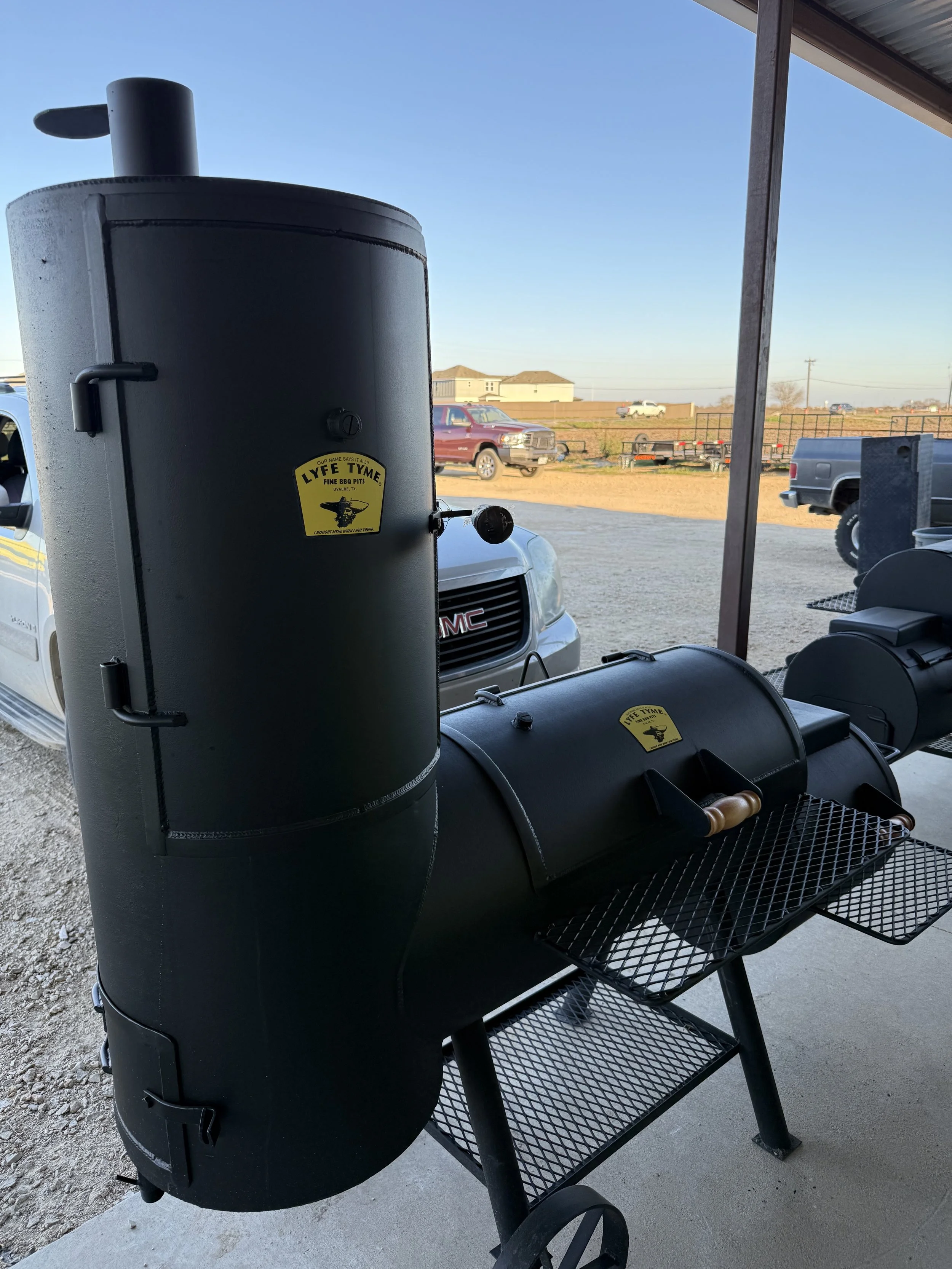 A black smoker grill with a chimney, sitting on a metal cart with wheels, located outside a building with a view of the parking lot and open field beyond.
