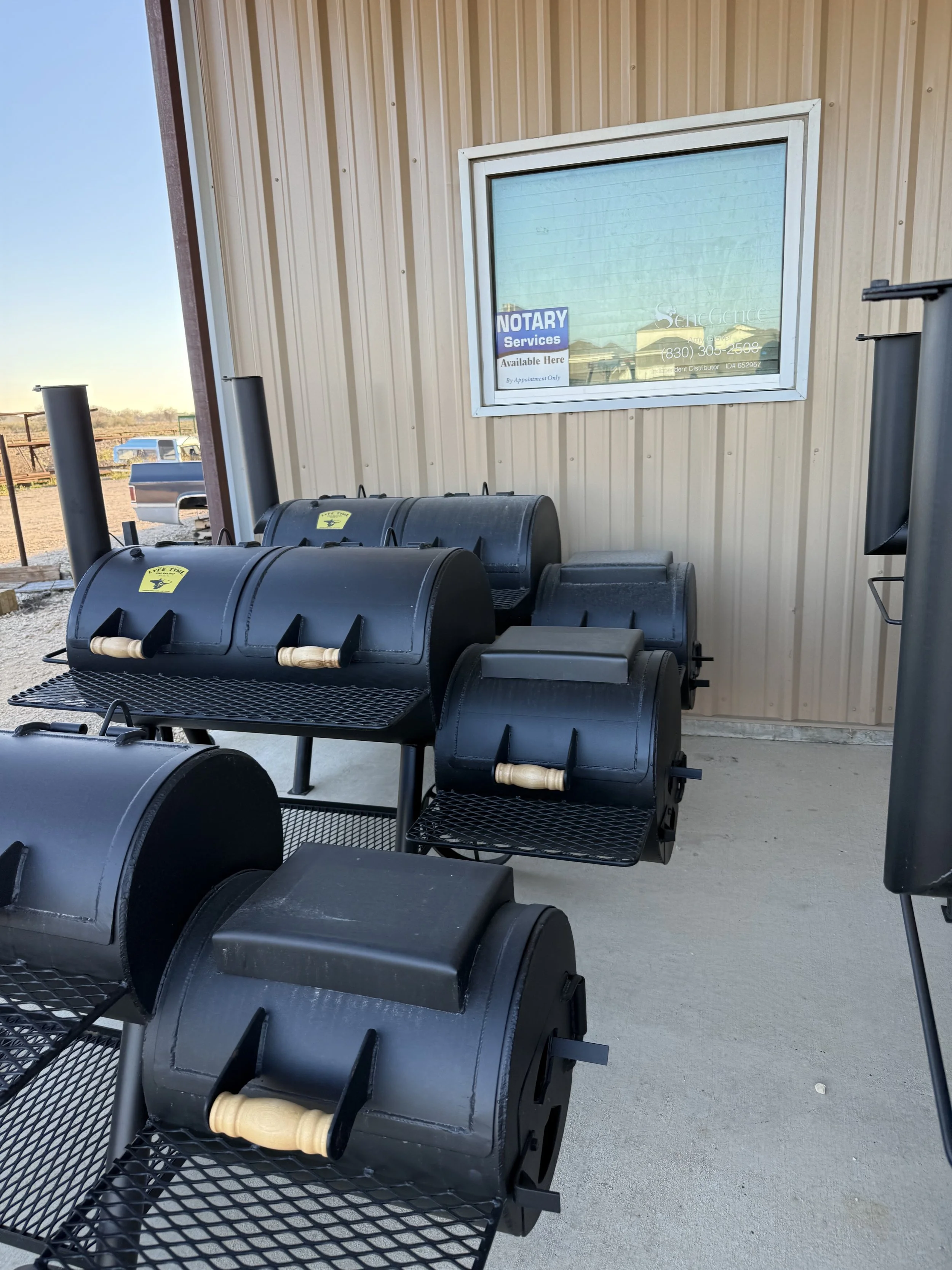 Several black outdoor smokers with wooden handles on a concrete patio against a beige metal building wall with a window that displays a sign reading 'Notary Services Available Here' and a reflection of the outdoor scene.