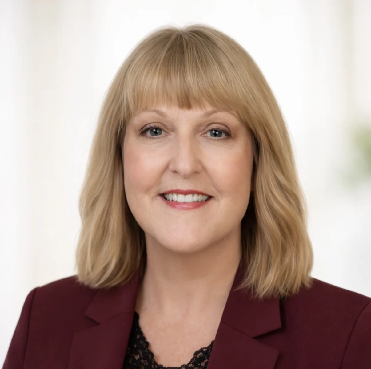 Portrait of a woman with shoulder-length blonde hair, wearing a maroon blazer and black lace top, smiling against a light, blurred background.