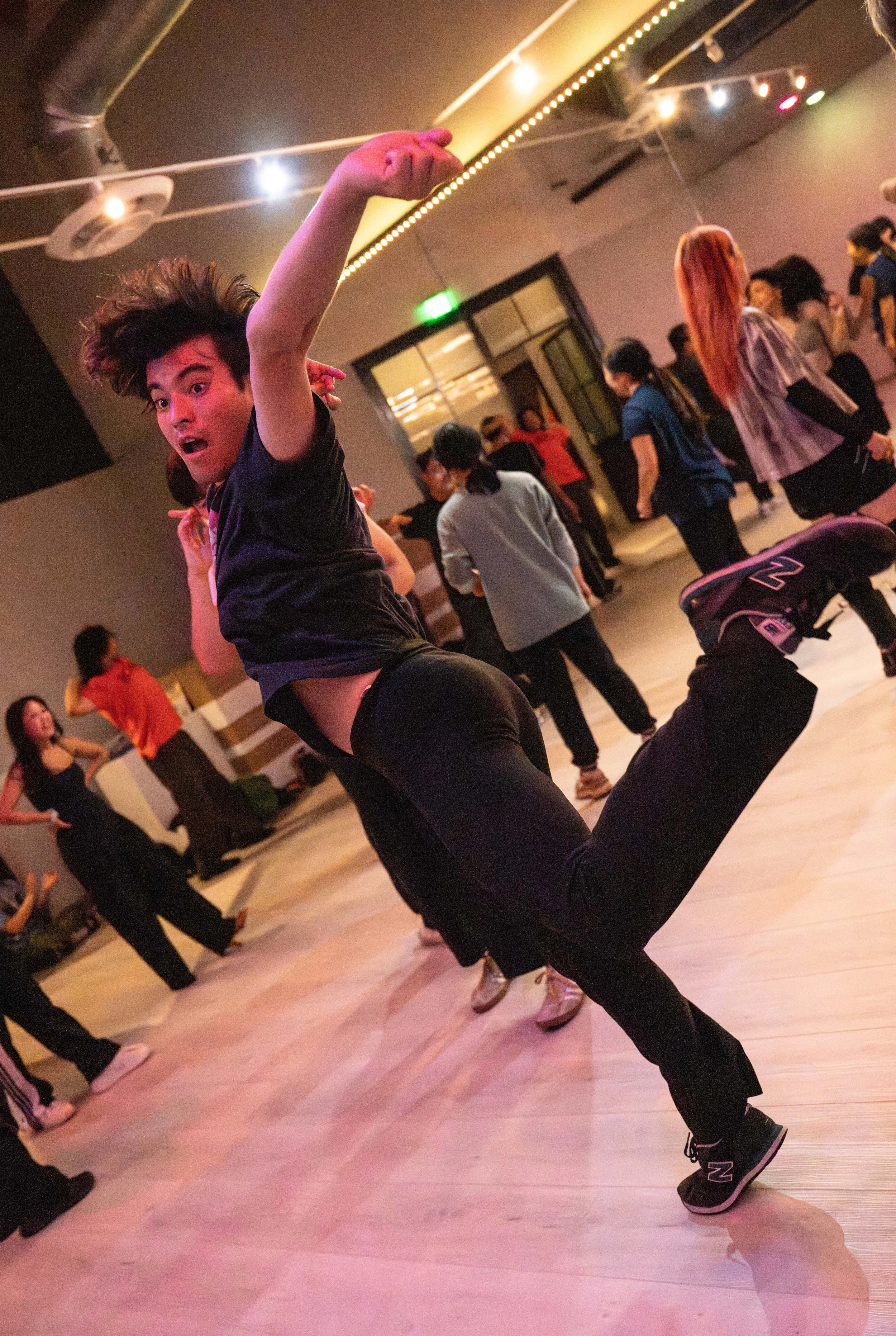 A person dancing in a lively dance studio with wooden floors and colorful lighting, surrounded by other dancers.