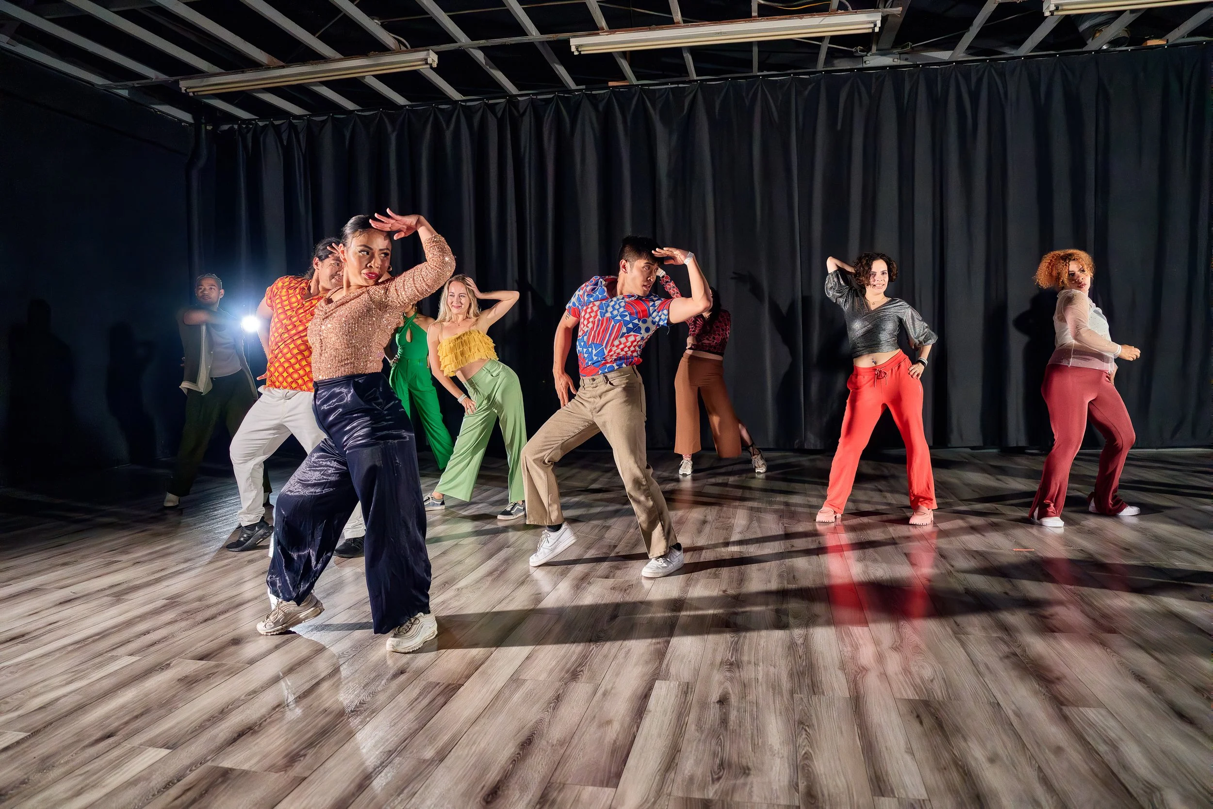 A group of diverse people dancing on a wooden floor in front of black curtains, with some people smiling and wearing colorful clothing.
