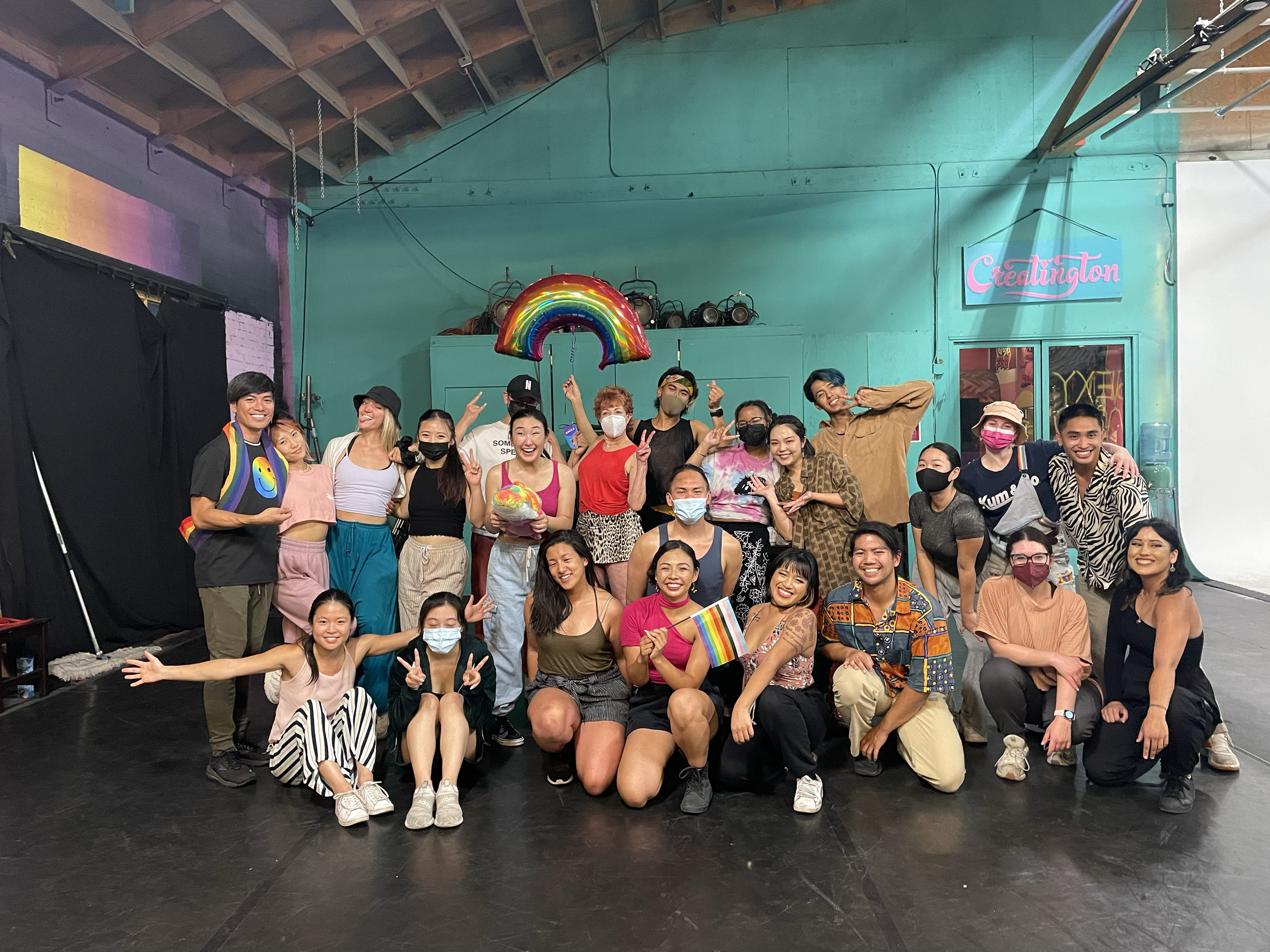 A diverse group of people celebrating at an indoor event, some holding rainbow flags and rainbow-themed props, with a rainbow balloon overhead. Many are smiling, with some making peace signs or other gestures. The setting has teal walls, and there is