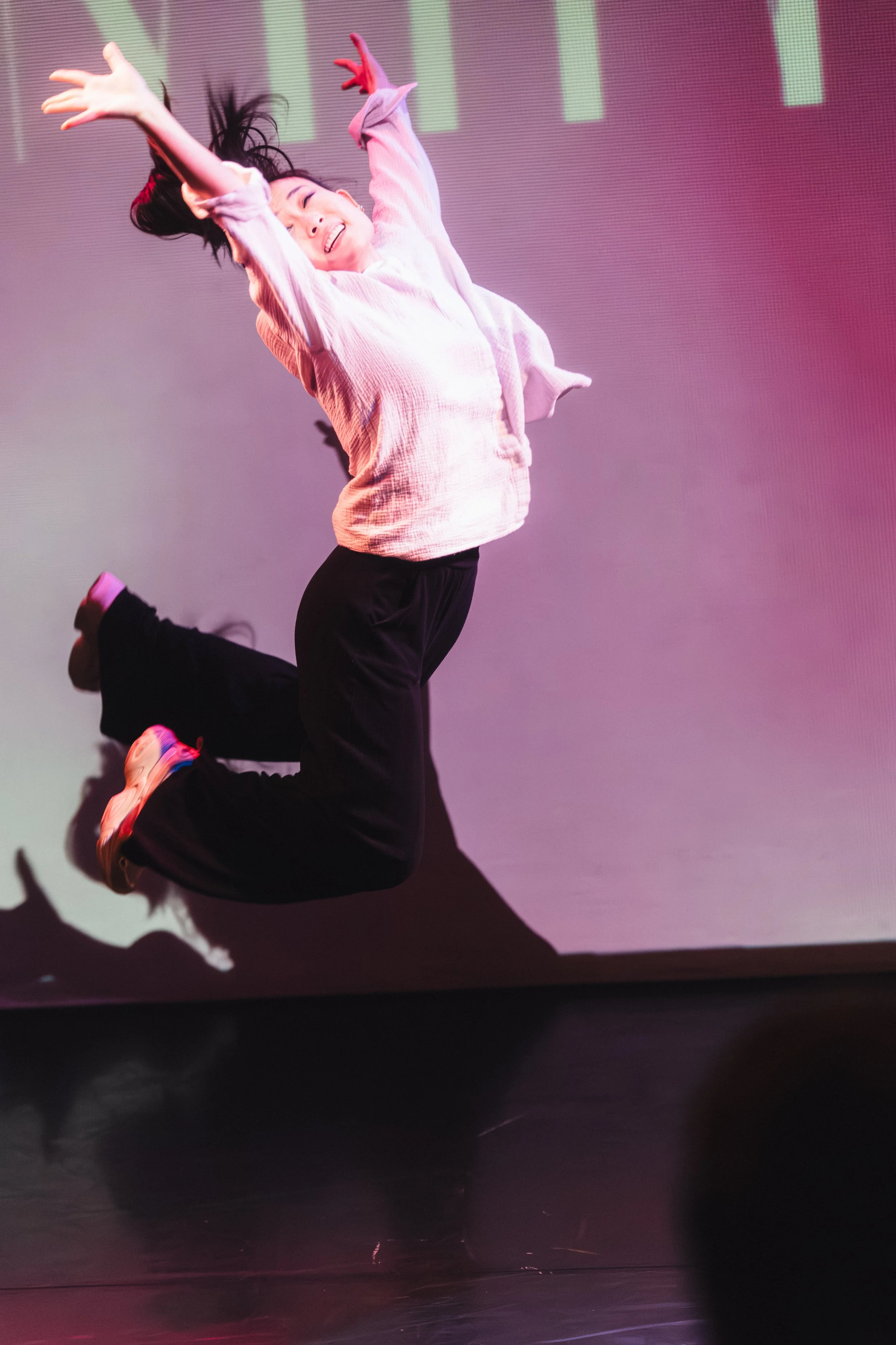 A woman is joyfully jumping in front of a colorful background during a live performance or event, wearing a light pink sweater and black pants.