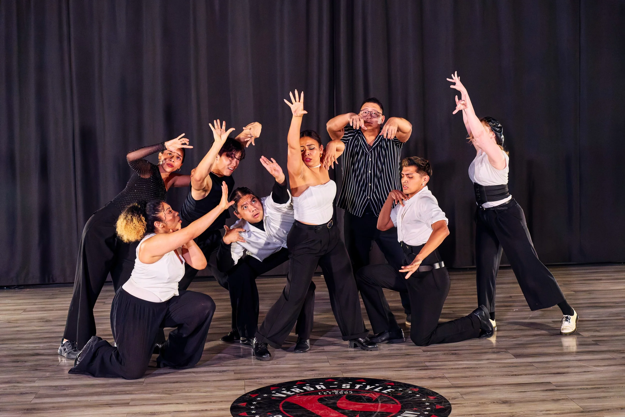 A group of nine diverse dancers performing on a stage with a dark curtain backdrop. They are in various expressive poses, some kneeling, some standing, and some reaching upward, dressed in black, white, and patterned clothing.