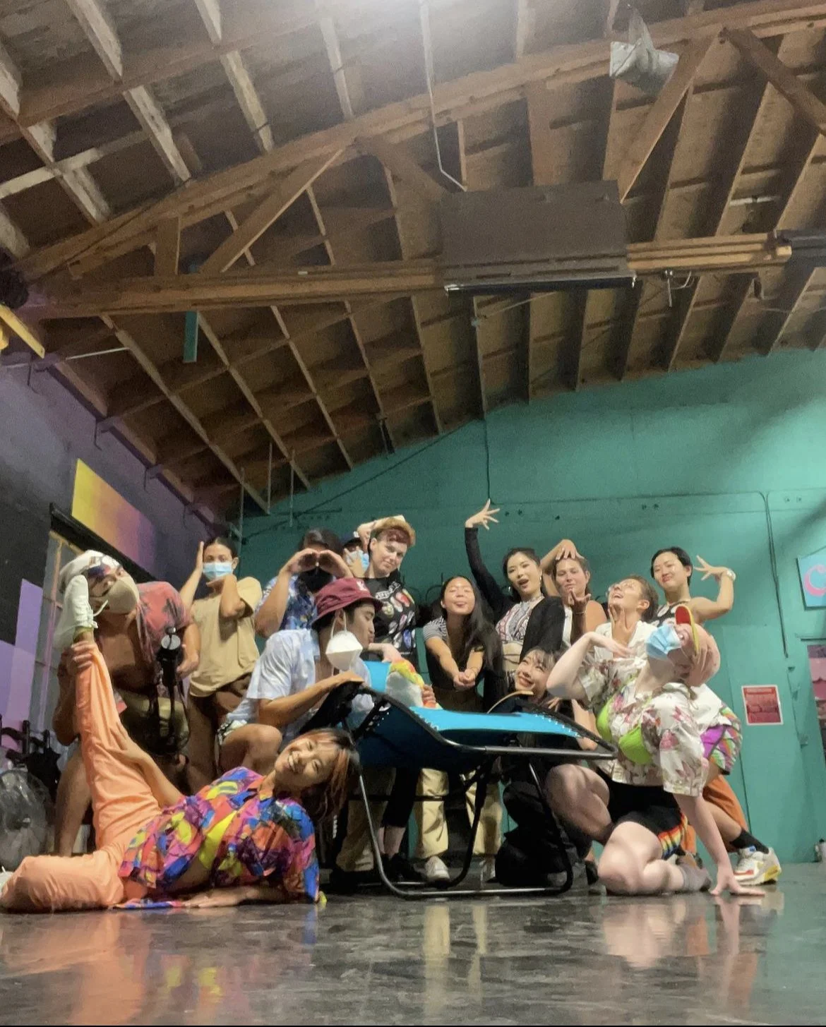 Group of diverse people, some wearing face masks, posing playfully in a studio with a green wall and wooden ceiling, some making peace signs, some sitting or lying on the floor.