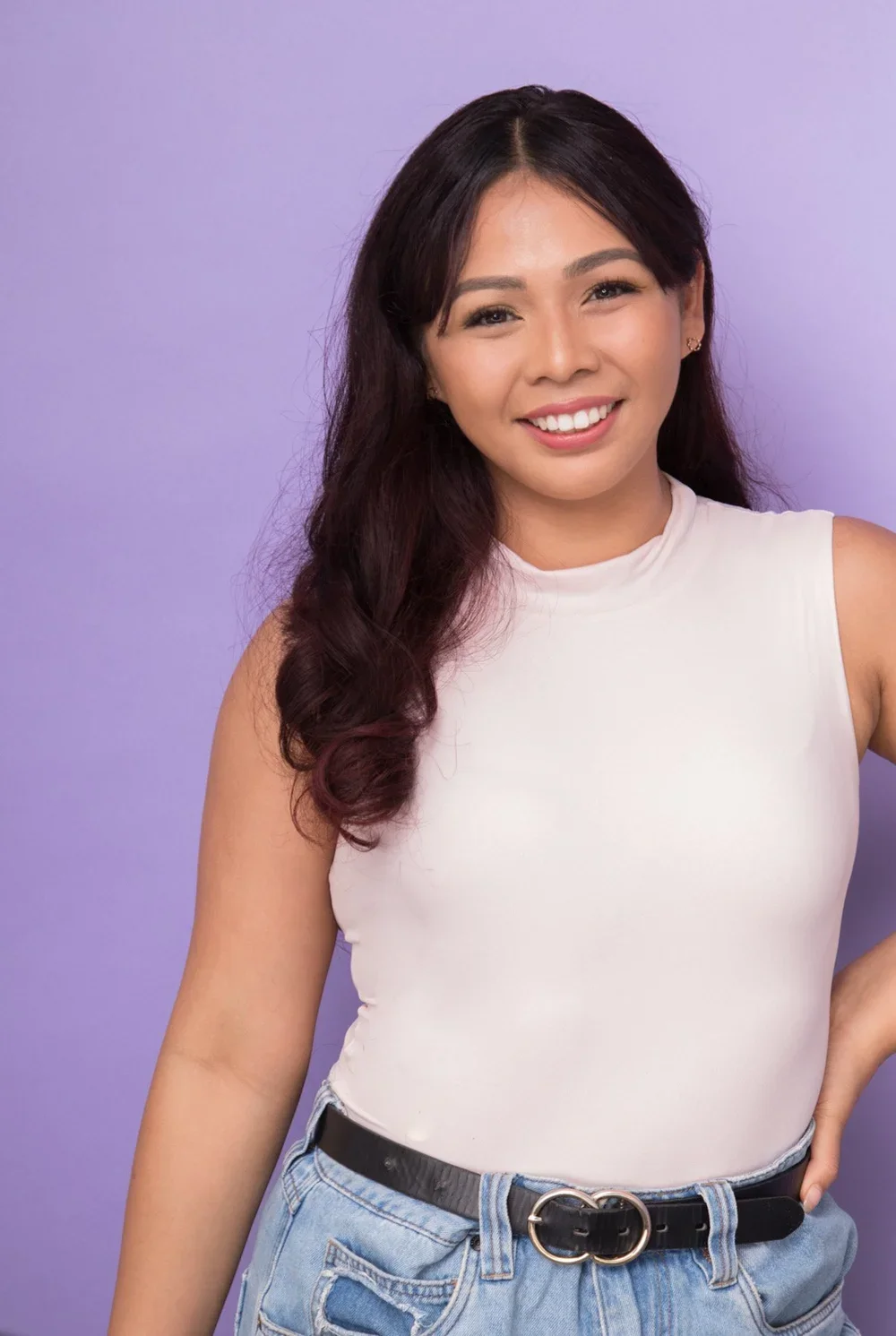 A young woman with long wavy dark hair smiling, wearing a white sleeveless top and light jeans with a black belt, standing against a purple background.