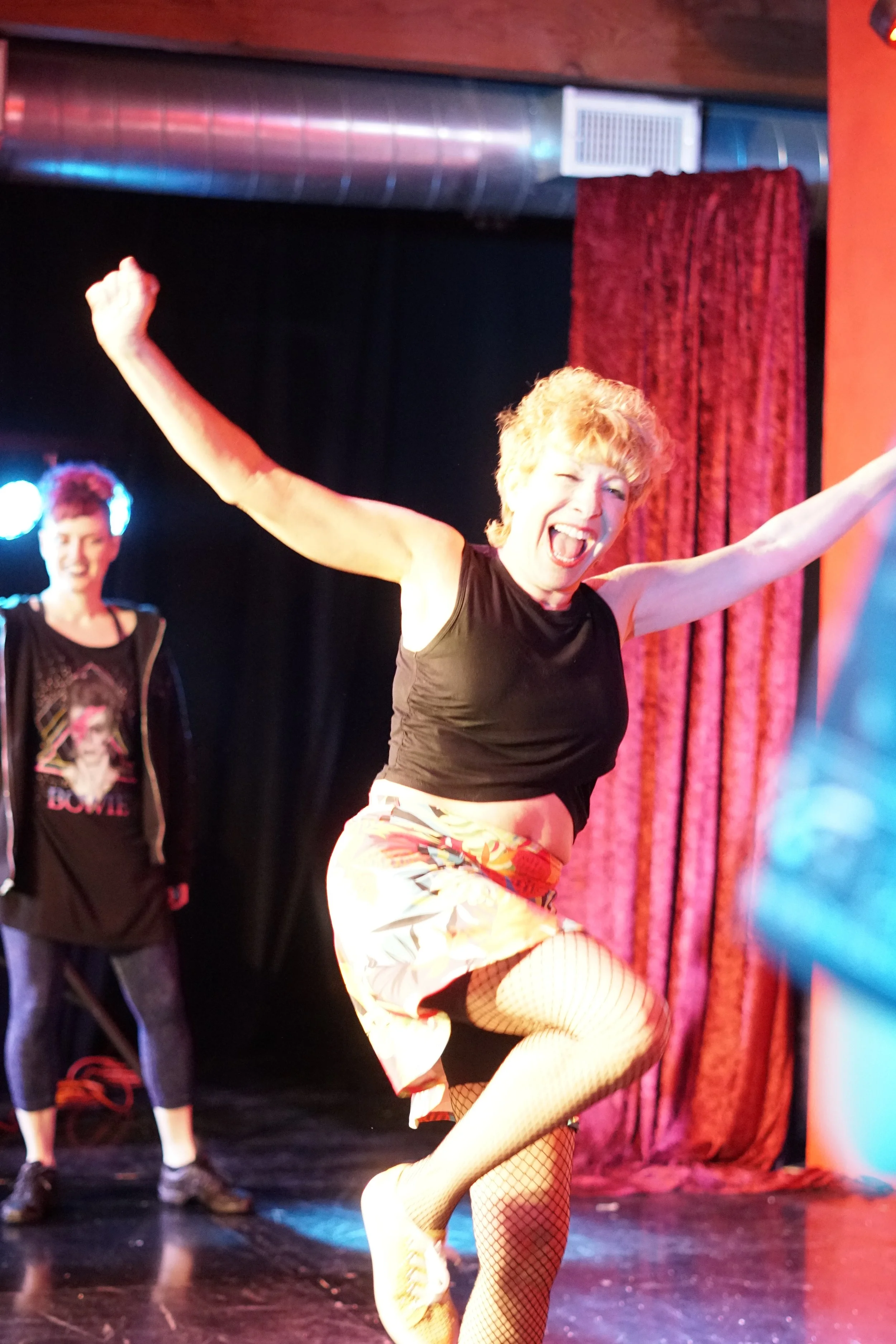 A woman with short blonde hair performing a dance move on stage, wearing a black sleeveless top, camo shorts, fishnet stockings, and lime green shoes. She is smiling and has her arms raised, with a red curtain in the background and another person sta