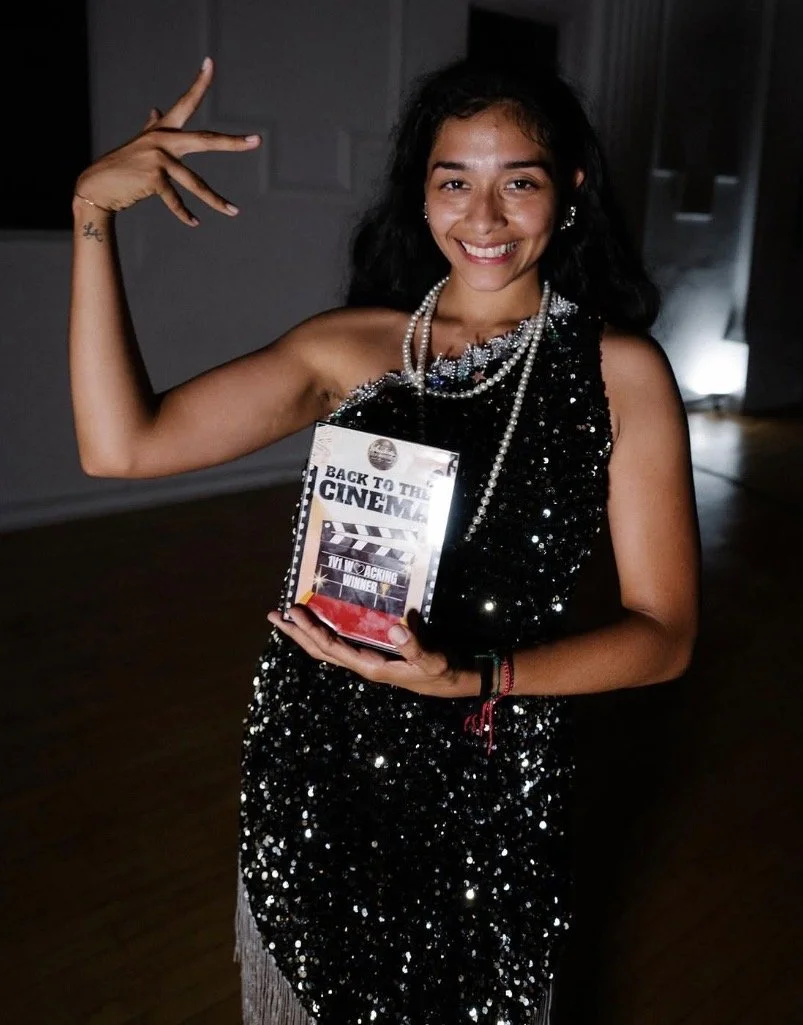 A woman with dark curly hair wearing a black sequined dress and pearl jewelry, smiling and holding a movie award or trophy, standing in a dimly lit room.