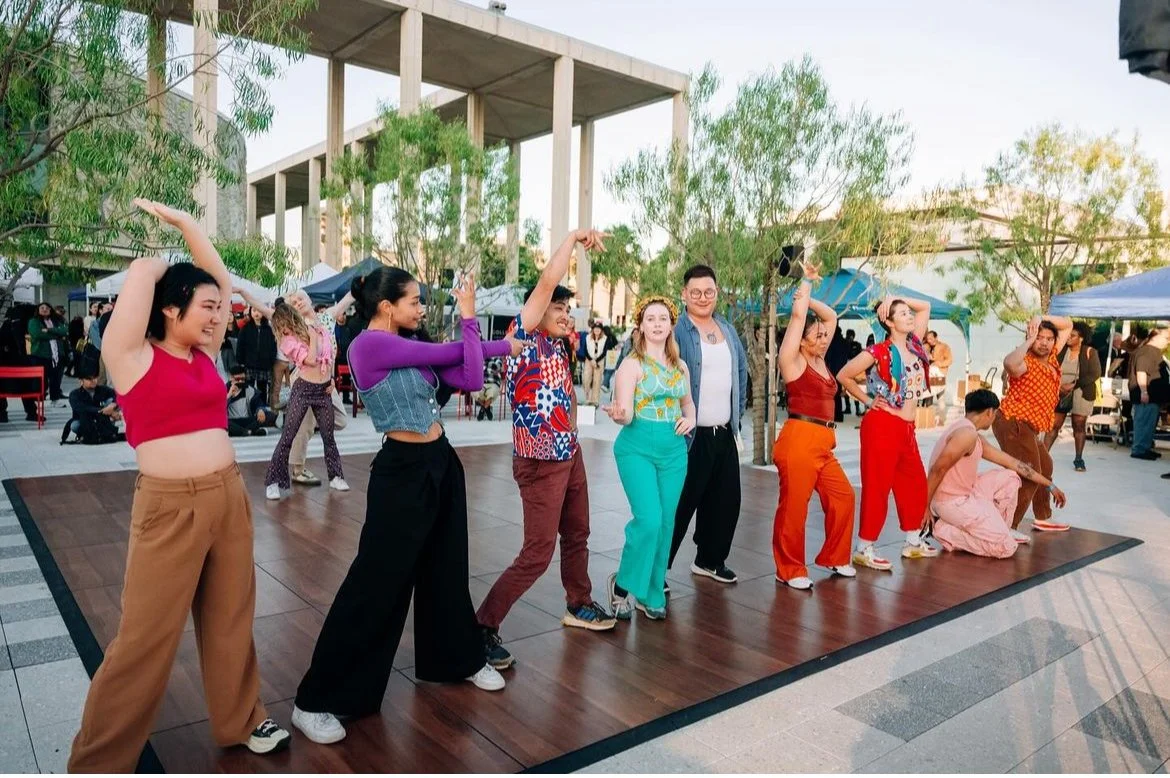 Group of people dancing on a wooden stage outdoors during daytime, with onlookers and trees in the background.