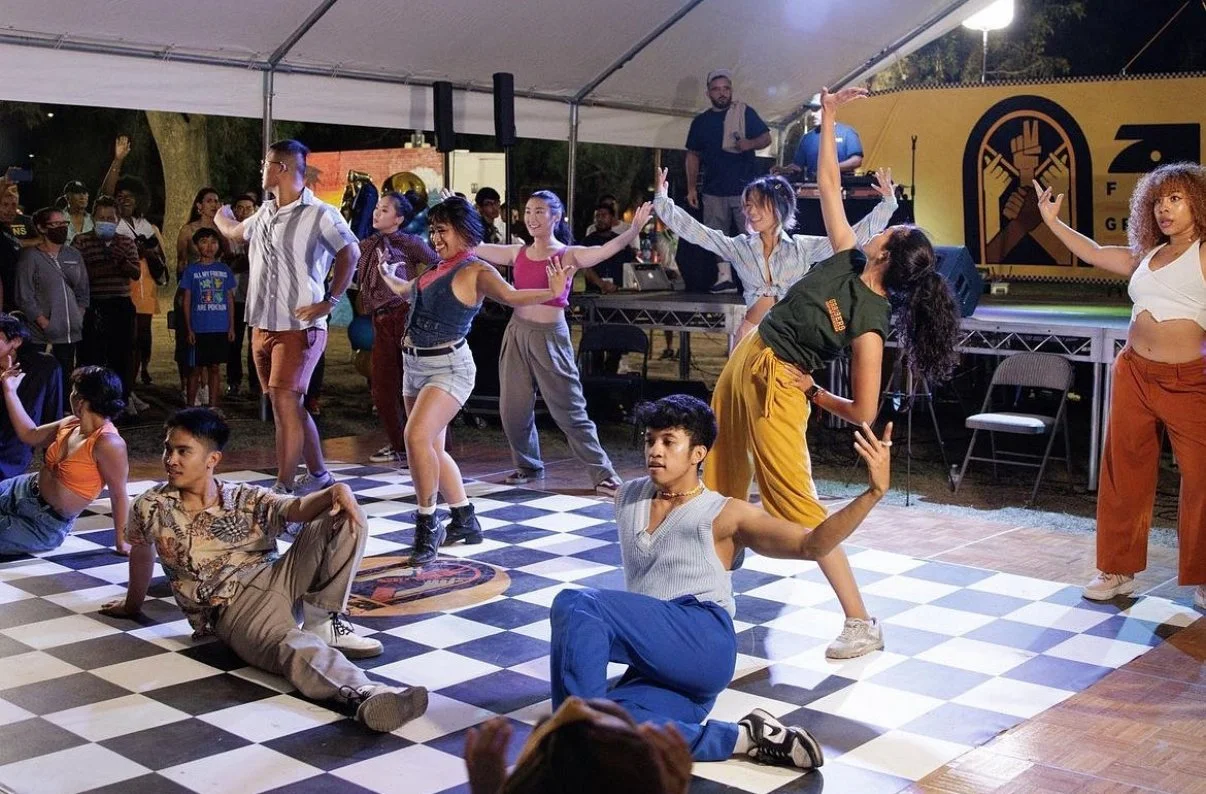 Group of young people dancing on a checkered floor at an outdoor event with a live DJ and audience watching.