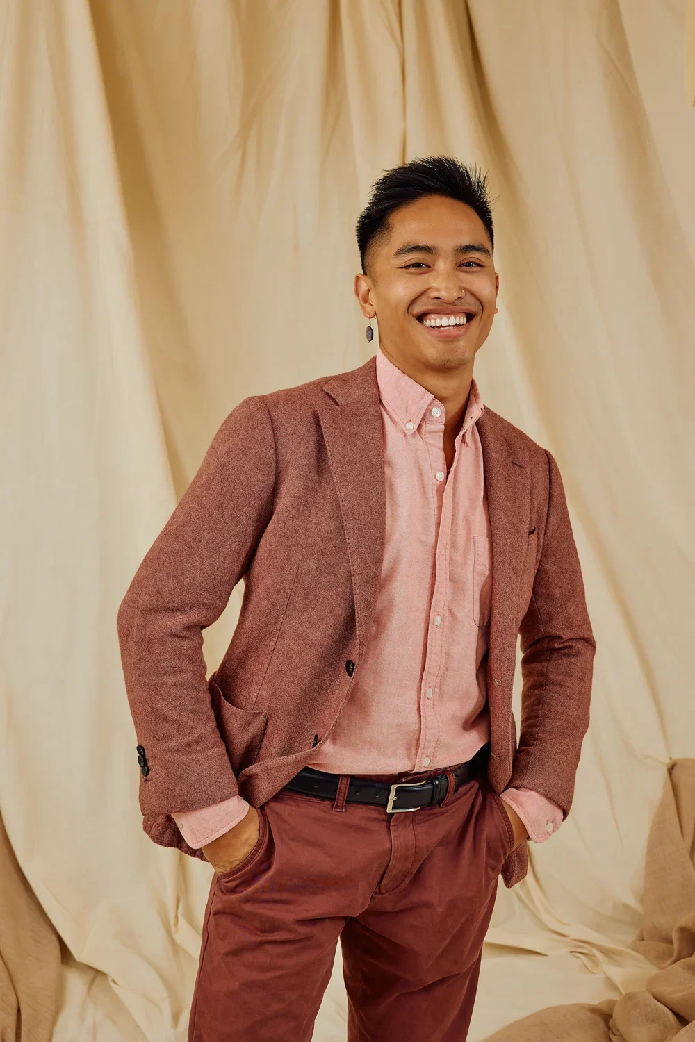A young man smiling, standing with hands in his pockets, wearing a pink shirt, a brown blazer, and burgundy pants, against a beige curtain backdrop.