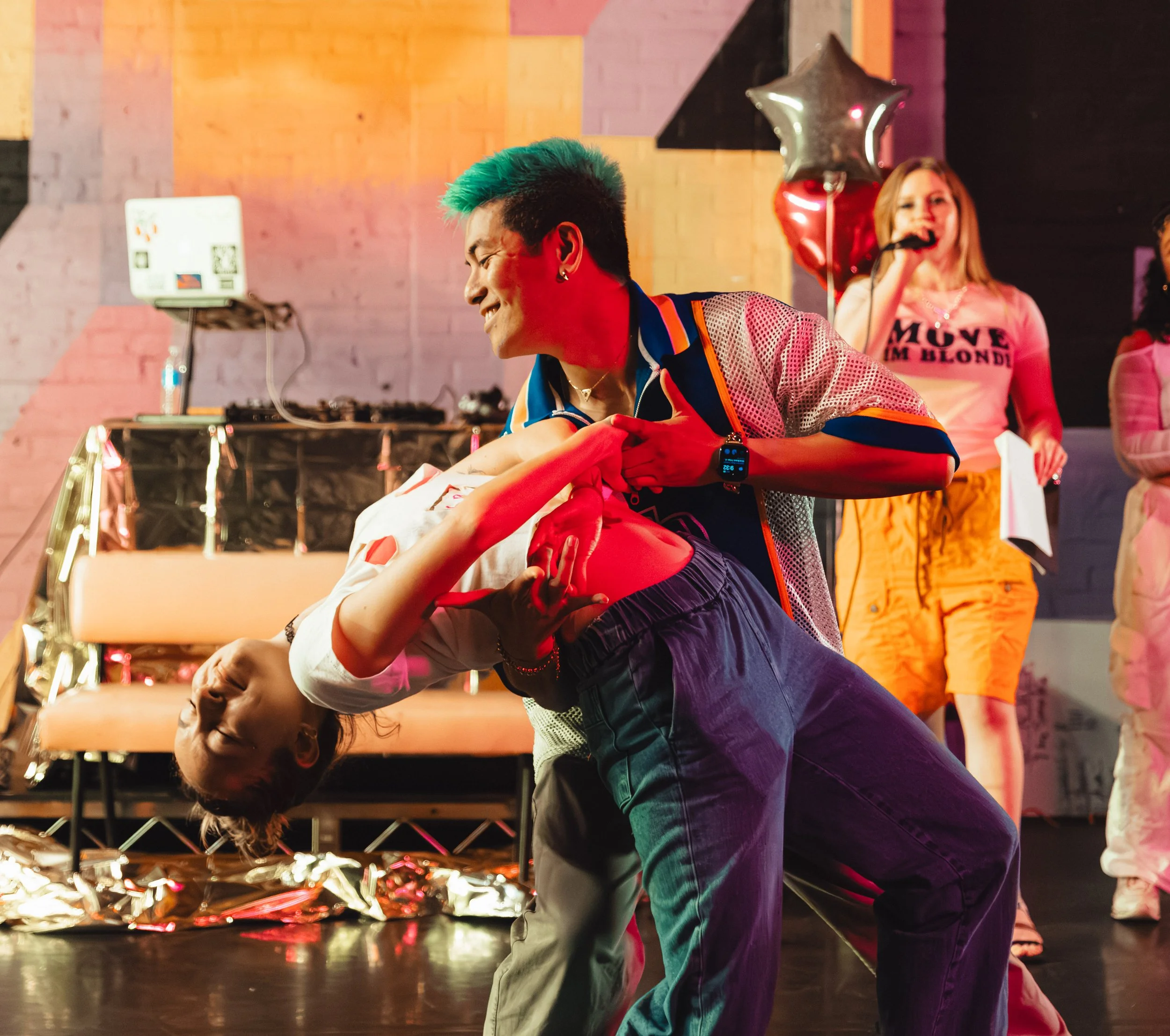 A man with teal-colored hair dancing with a woman in a dark setting decorated with balloons and star-shaped balloons. The man is smiling and appears to be enjoying himself. In the background, a woman is holding a microphone, indicating a celebration or party atmosphere.