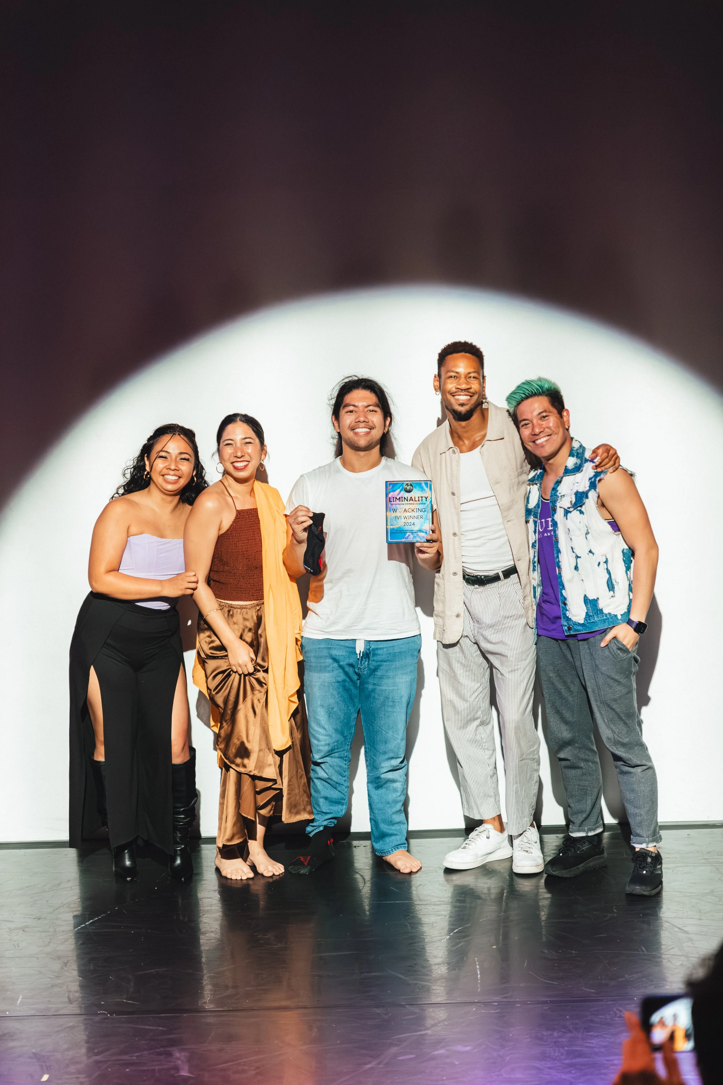 A diverse group of six people standing on stage, smiling, with one holding an award plaque that says 'Liminality. Tackling Myths Winner 2024'. The group includes men and women of different ethnicities, some barefoot and some in shoes, dressed in casu