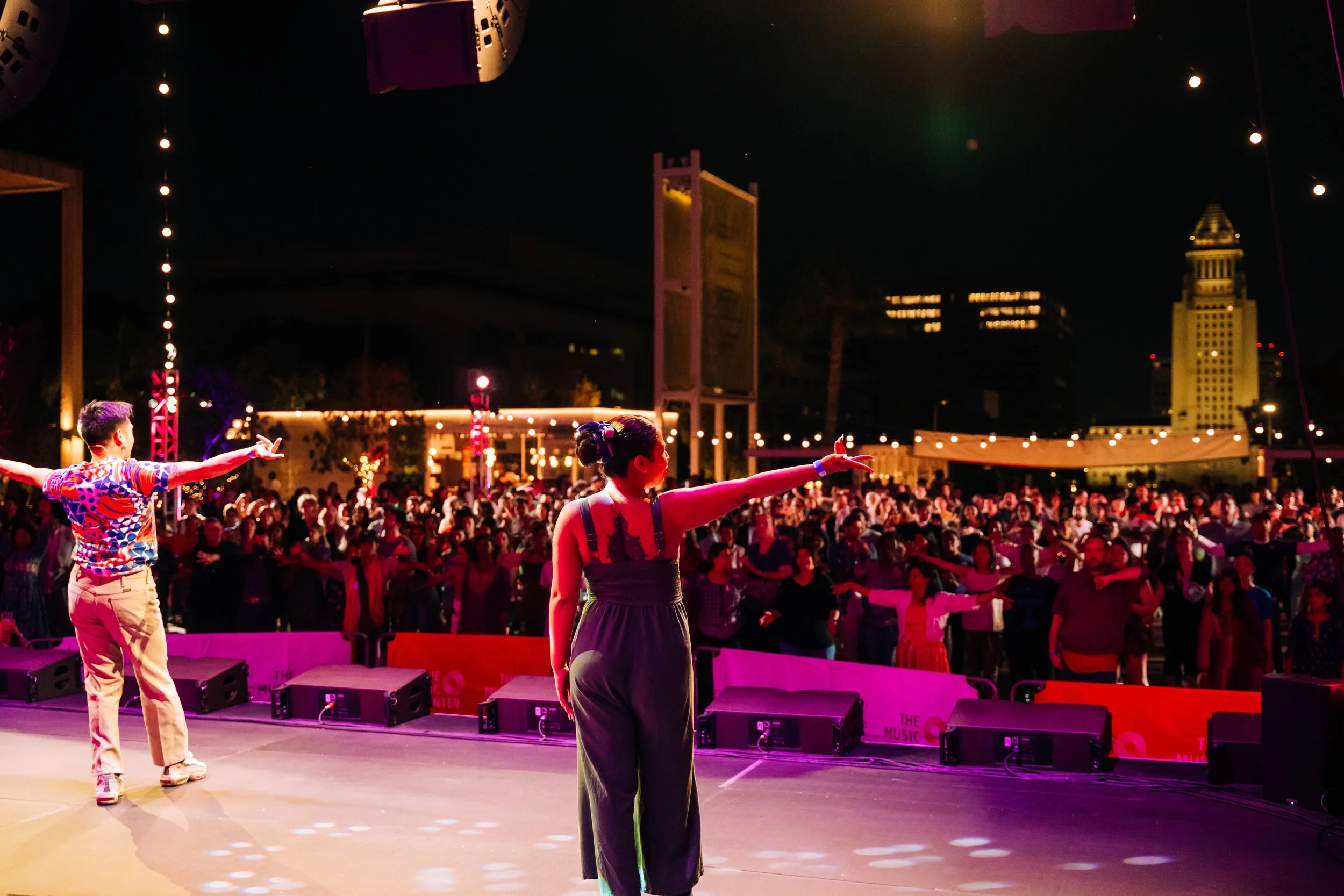 A stage performance with dancers in front of a large audience at night, city skyline with tall buildings and a clock tower visible in the background, festive lights overhead.