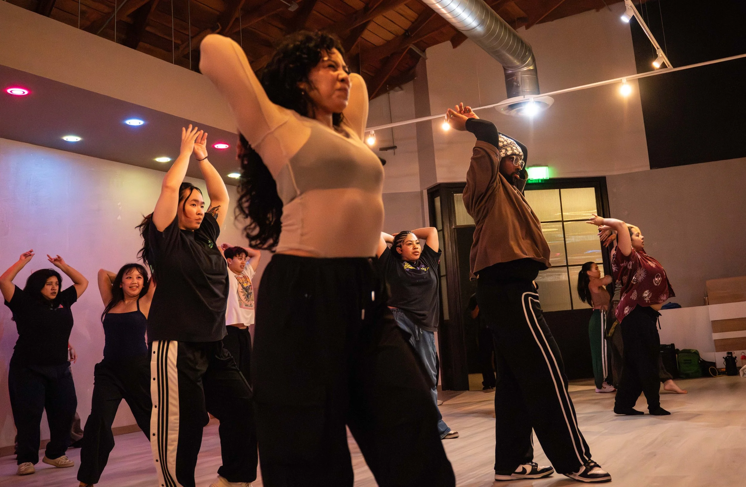 Group of diverse people practicing dance or yoga in a studio with a wooden ceiling and colorful lights.