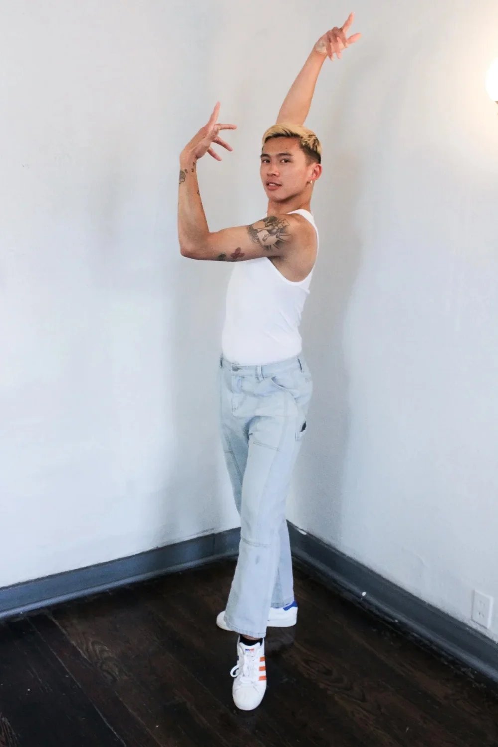Person with tattoos in a dance pose, wearing a white tank top, light-colored jeans, and white sneakers with orange stripes, standing in a corner of a room with dark hardwood floors and plain white walls.
