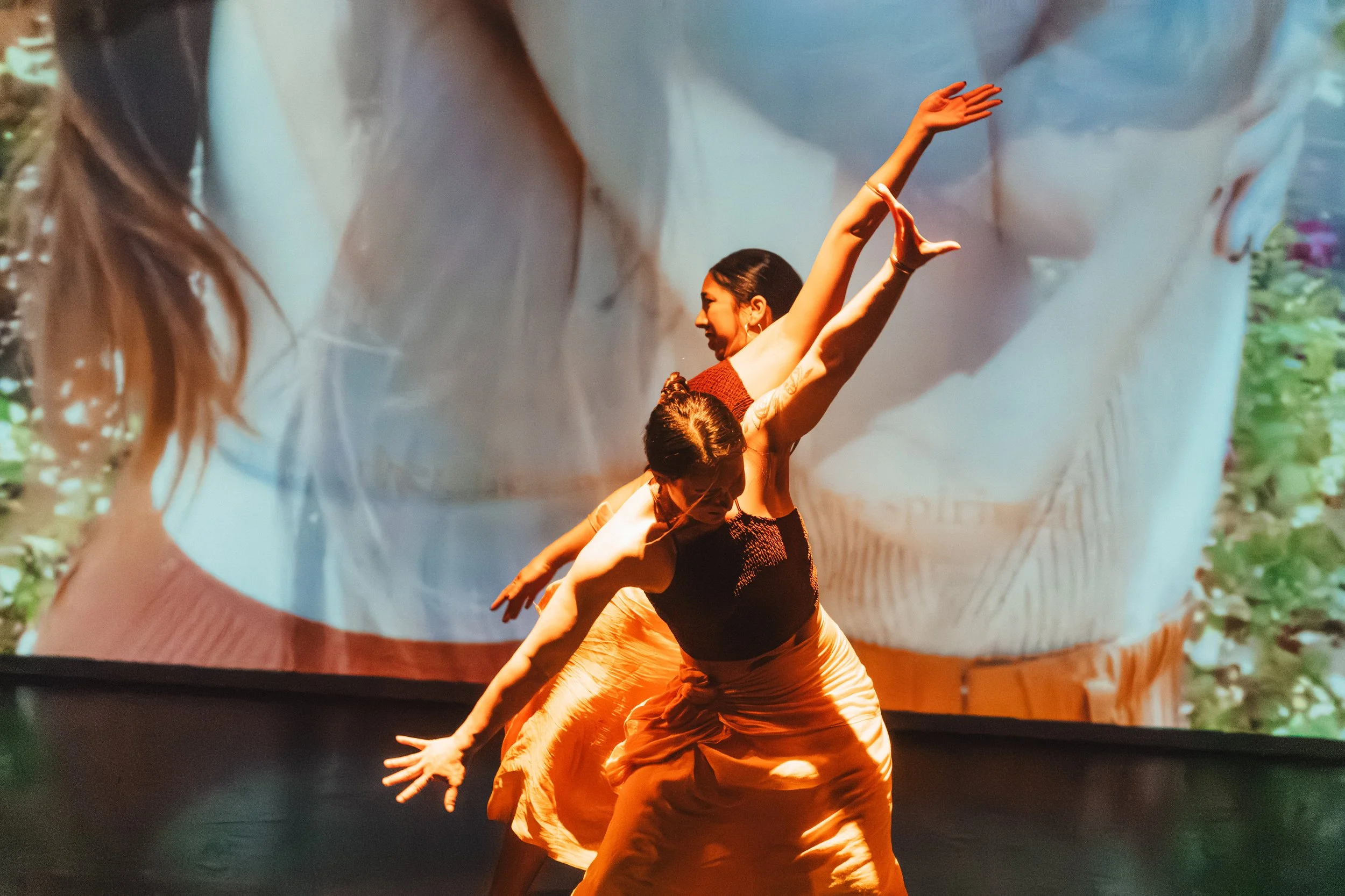 A woman dancing on stage, wearing a black top and orange skirt. Her arms are raised in the air and she appears to be performing in front of a colorful backdrop.