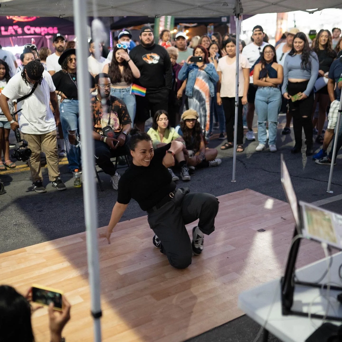 A woman with short dark hair and black clothing is crouching and smiling on a wooden dance floor at an outdoor event, with a crowd of onlookers behind her under tents at dusk.