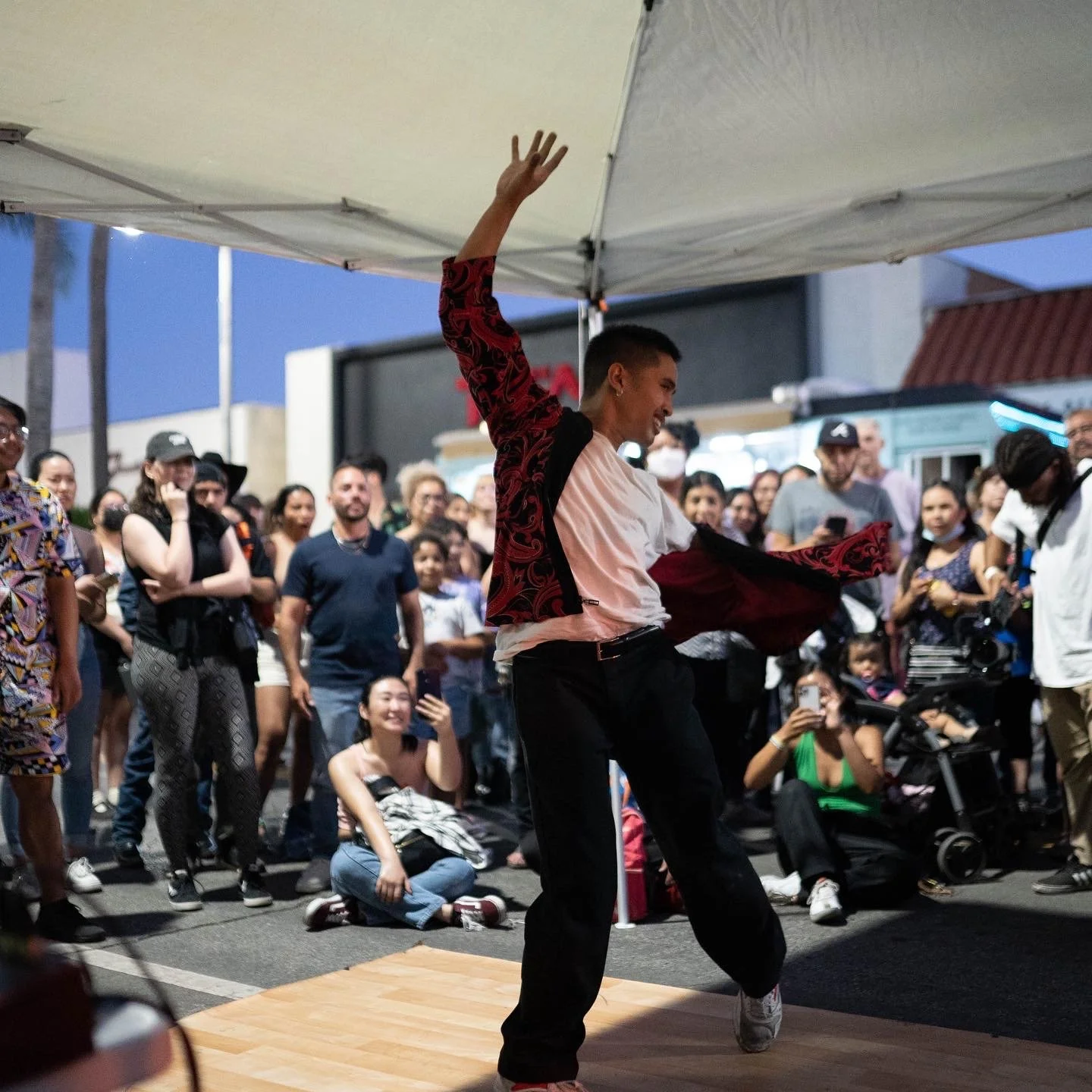 A dancer performing on a small wooden stage in front of a crowd gathered outside during the evening. The dancer is wearing a white t-shirt and black pants with a red and black patterned jacket, and is in a pose with one arm raised and a smile on his 