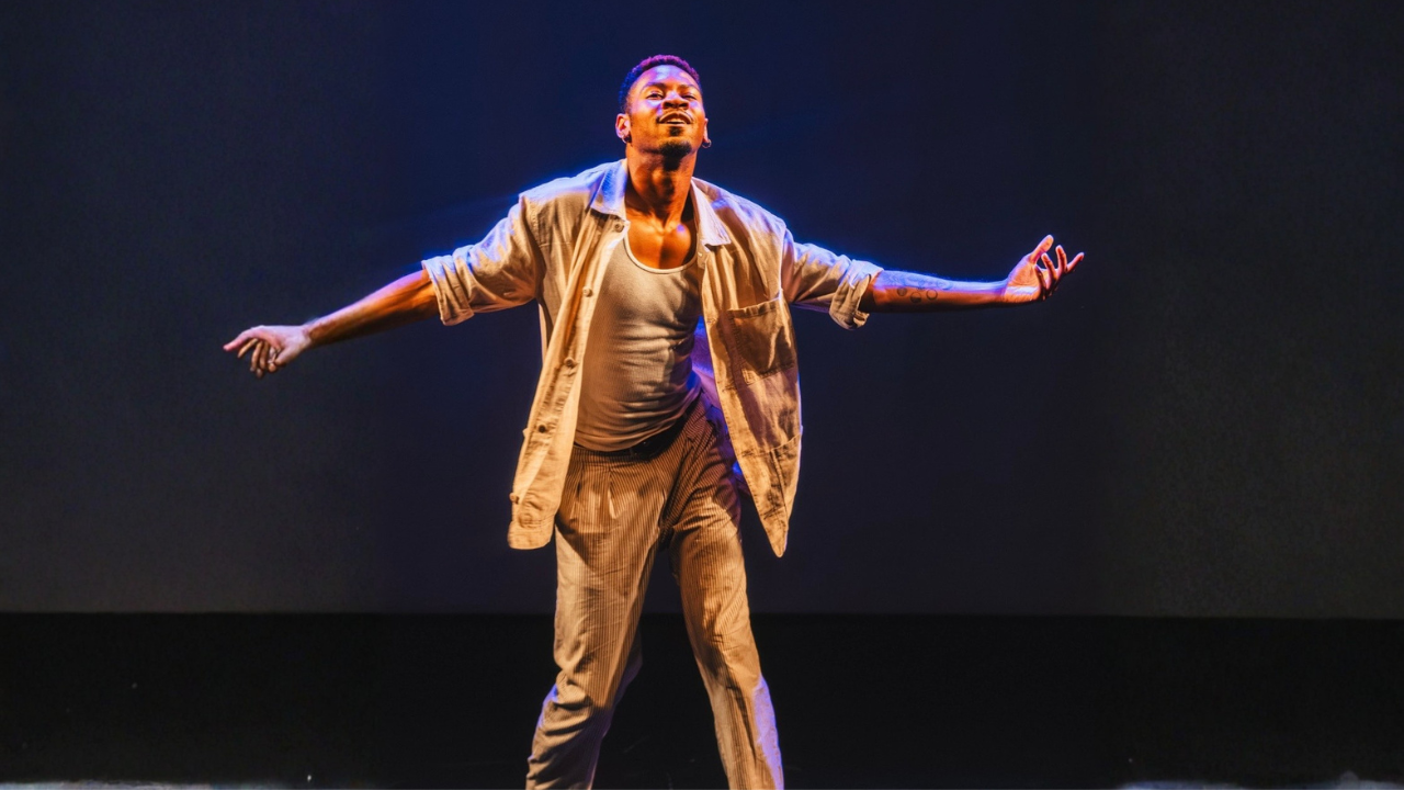 Performer on stage with arms outstretched, facing upward, wearing casual clothing and expressive stance