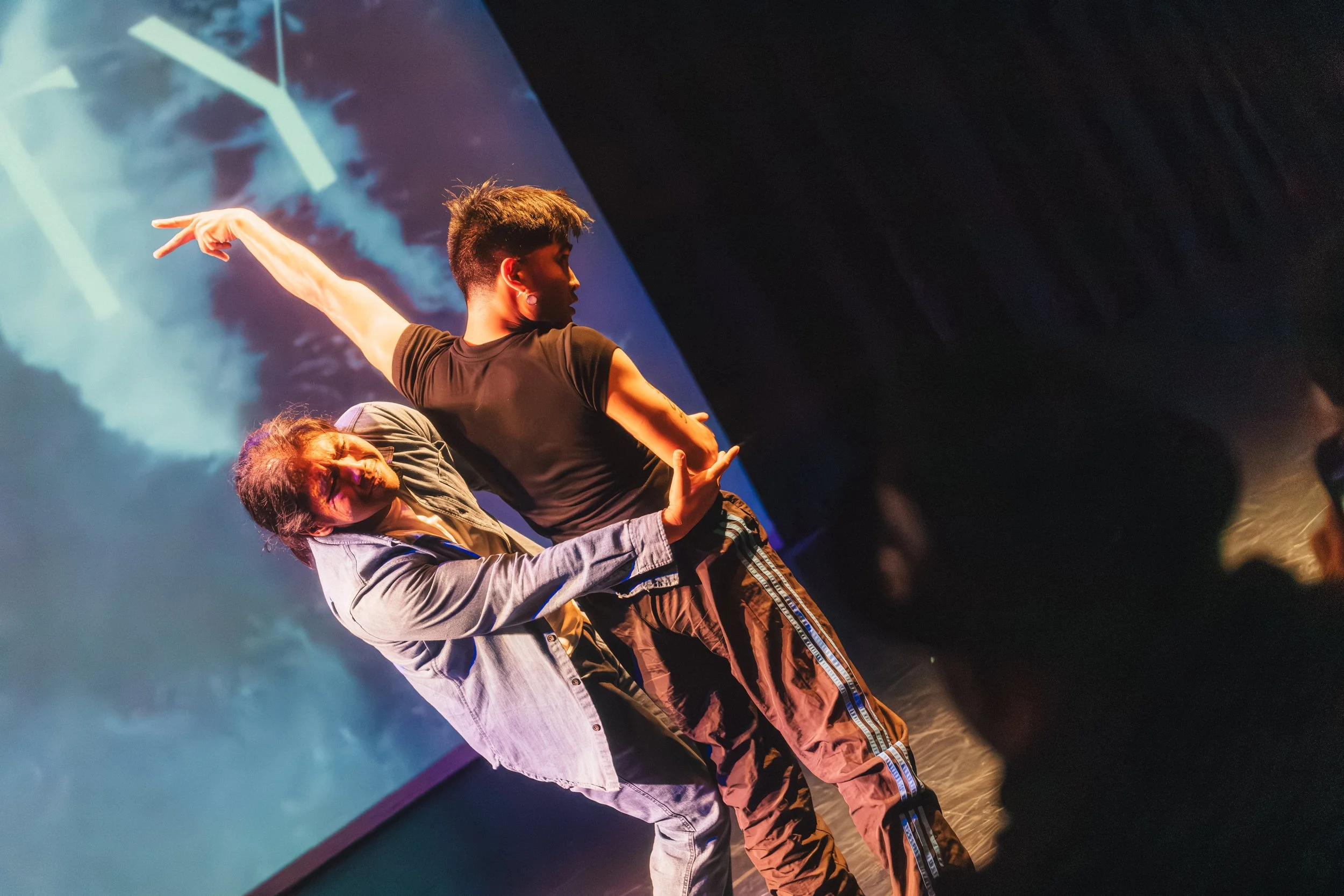 Two dancers perform on a stage with blue lighting and abstract background projection.