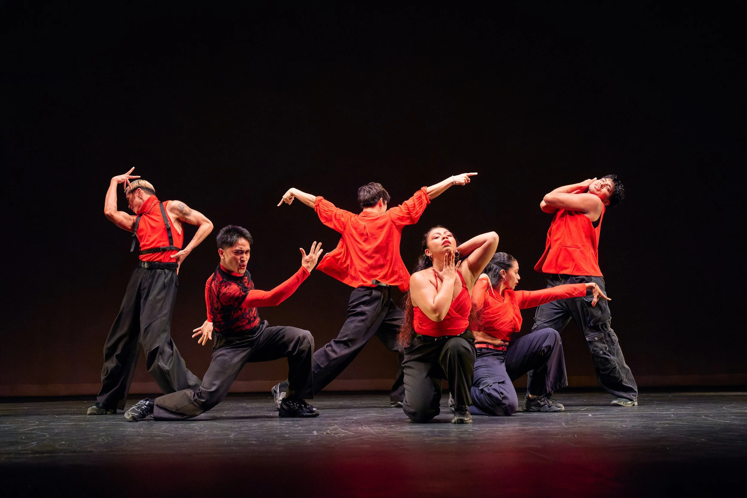 Dancers on stage wearing red and black costumes, performing a theatrical dance routine against a black background.