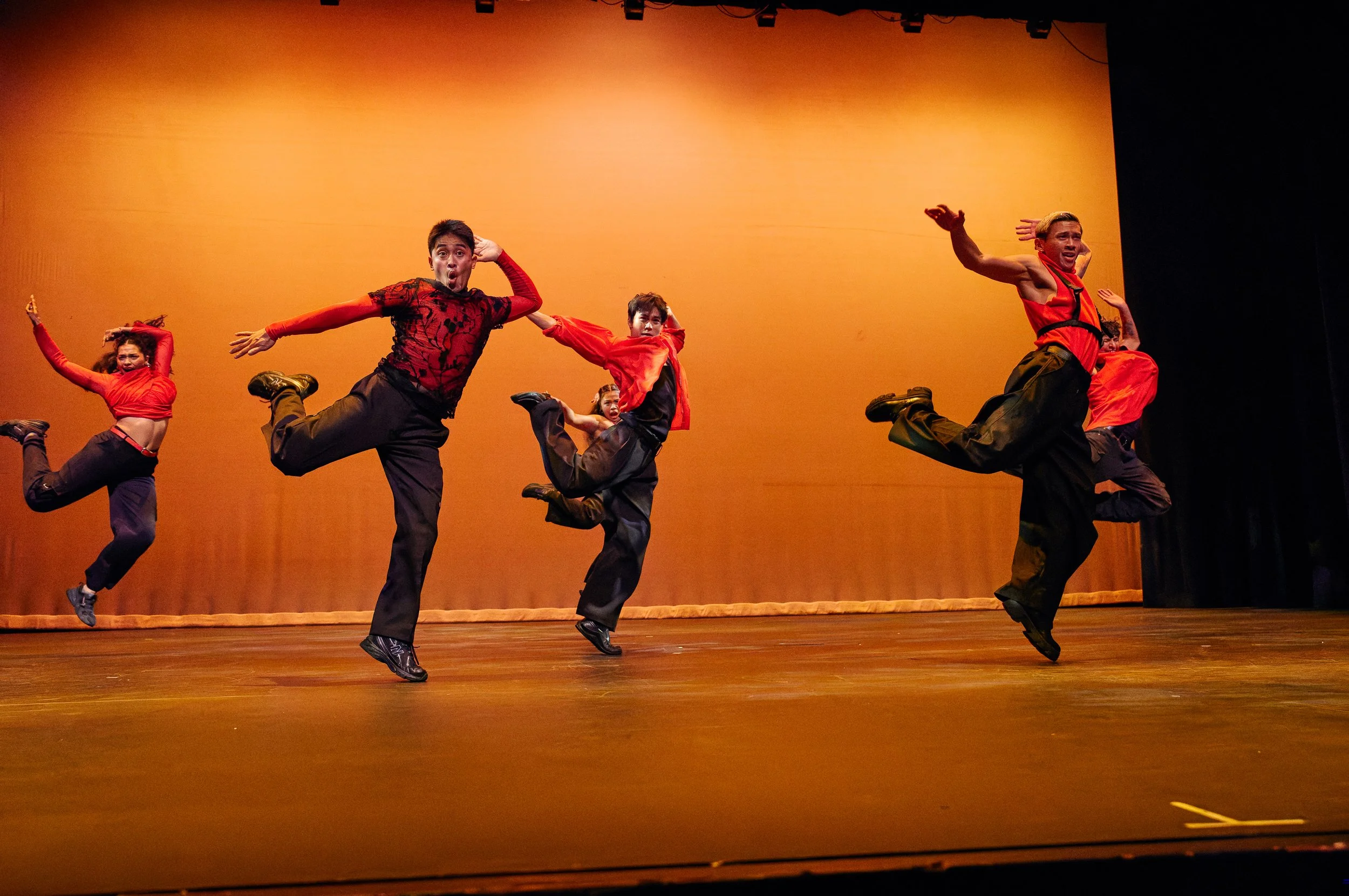 Dance performance on stage featuring dancers in red and black costumes with energetic poses against an orange background.