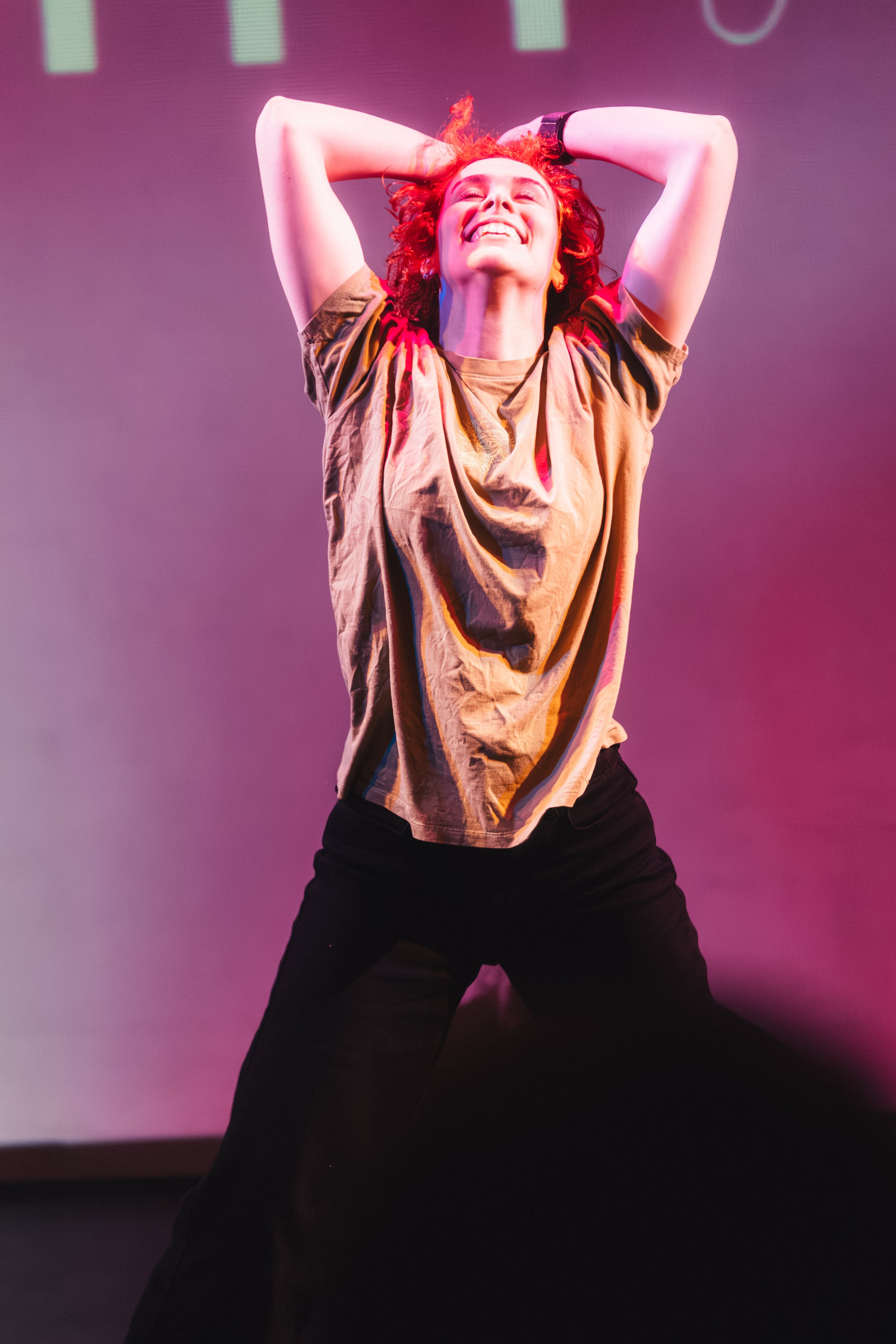 A woman with curly red hair smiling with her eyes closed and arms raised, wearing a tan shirt and black pants, in front of a purple background.