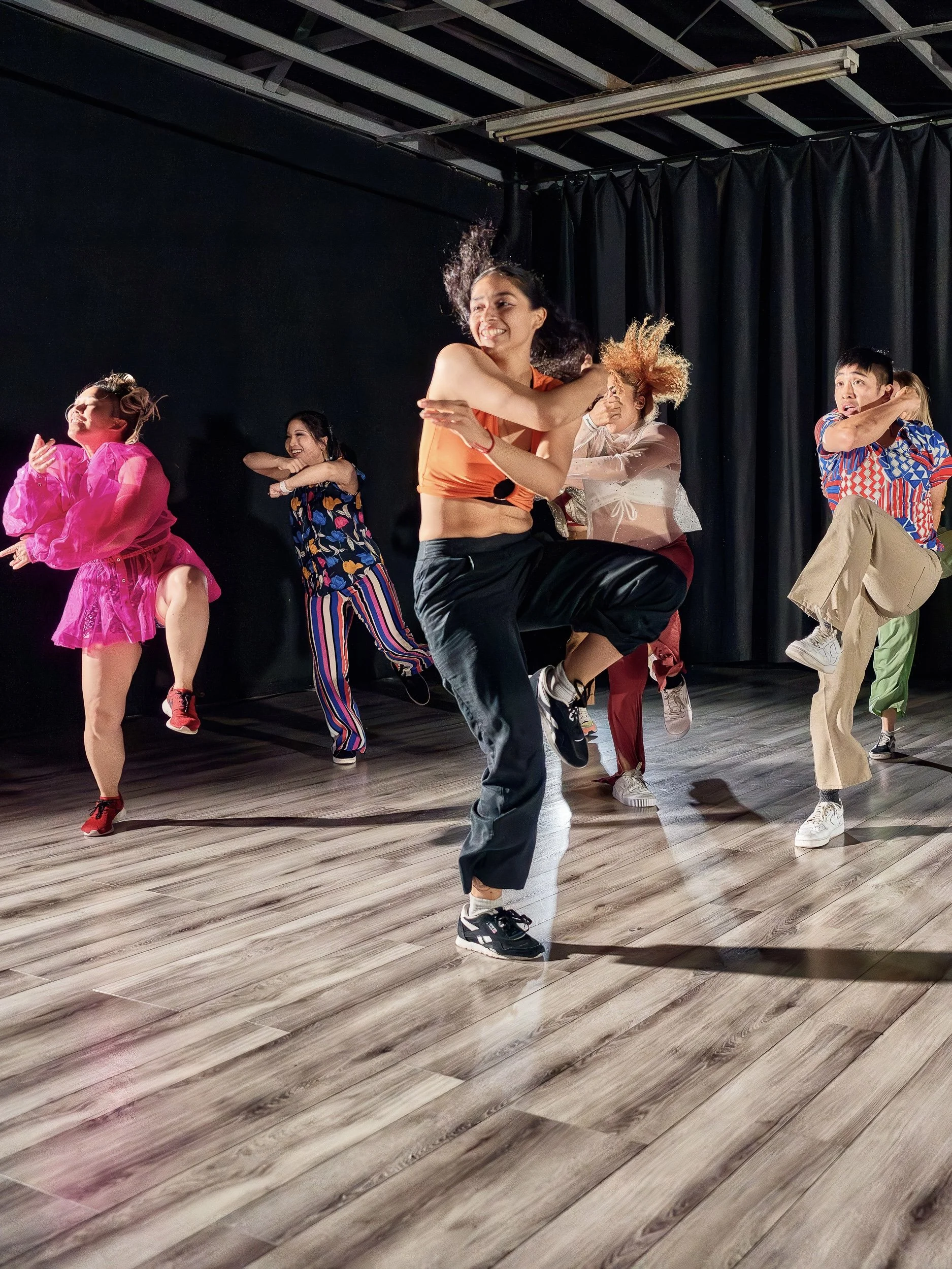 A group of diverse young dancers performing a lively dance routine on a stage with black curtains. They are wearing colorful, casual outfits and showing energetic movement and expressions.