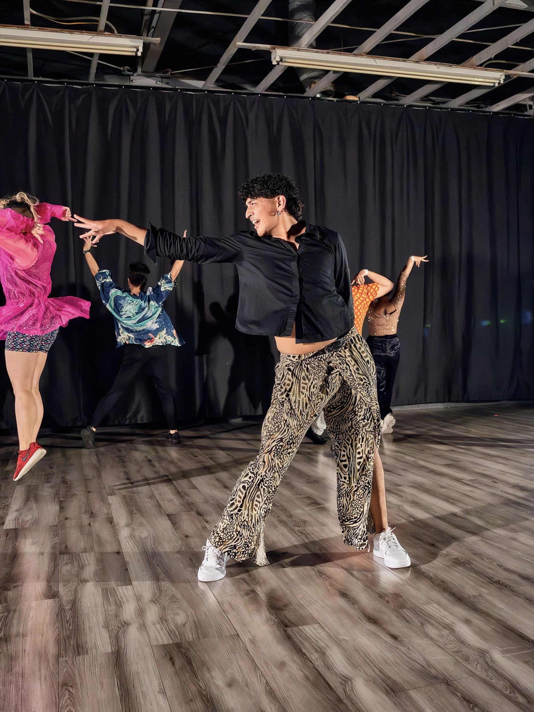 A group of people dancing and performing on a stage with black curtains in the background. The person in the center is wearing animal print pants and a black shirt, striking a pose. The others are in colorful outfits with vibrant patterns and are mid