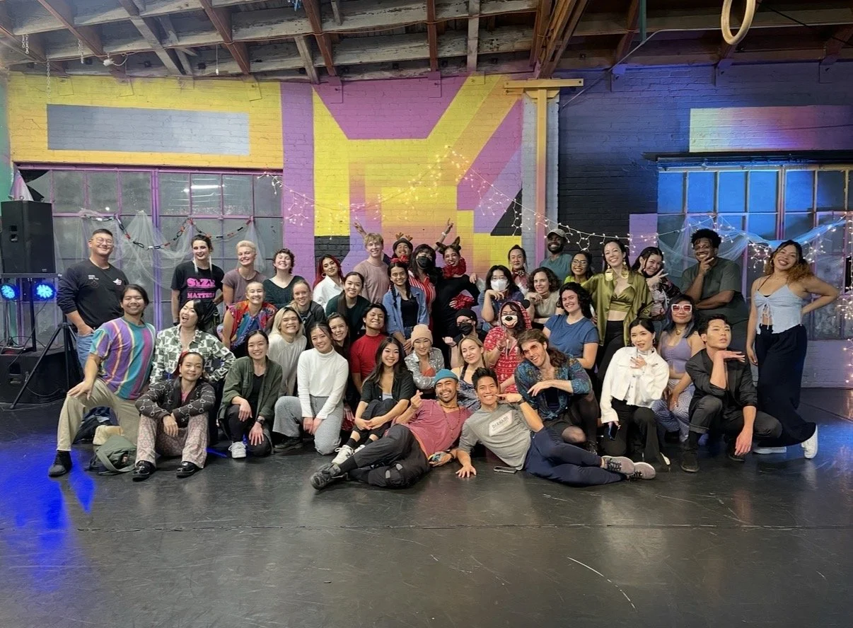 A diverse group of people posing together in a colorful, decorated indoor space with bricks on the wall and string lights, celebrating at an event or party.