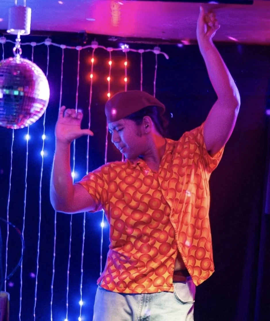 A person dancing with one arm raised and eyes closed in a colorful, neon-lit setting with a disco ball and string lights in the background.