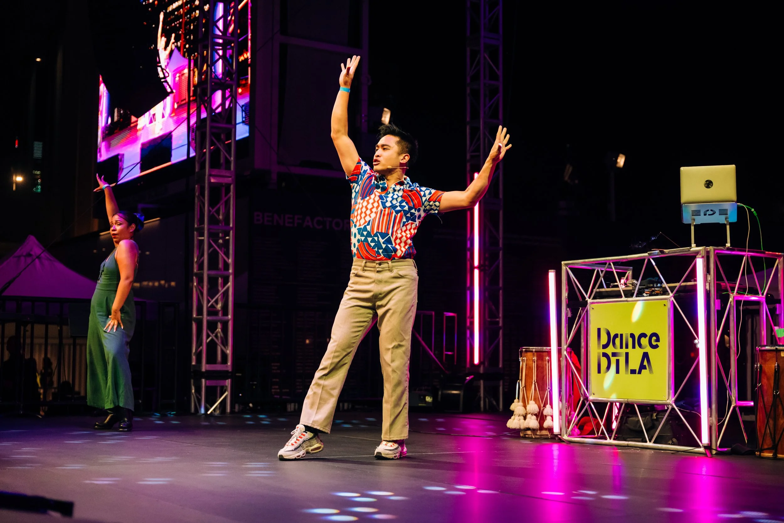 A male performer with short black hair wearing a colorful patterned shirt, khaki pants, and sneakers dancing on stage at an event called 'DanceATL,' with a woman in a green dress in the background and a DJ setup with a yellow laptop on the right.