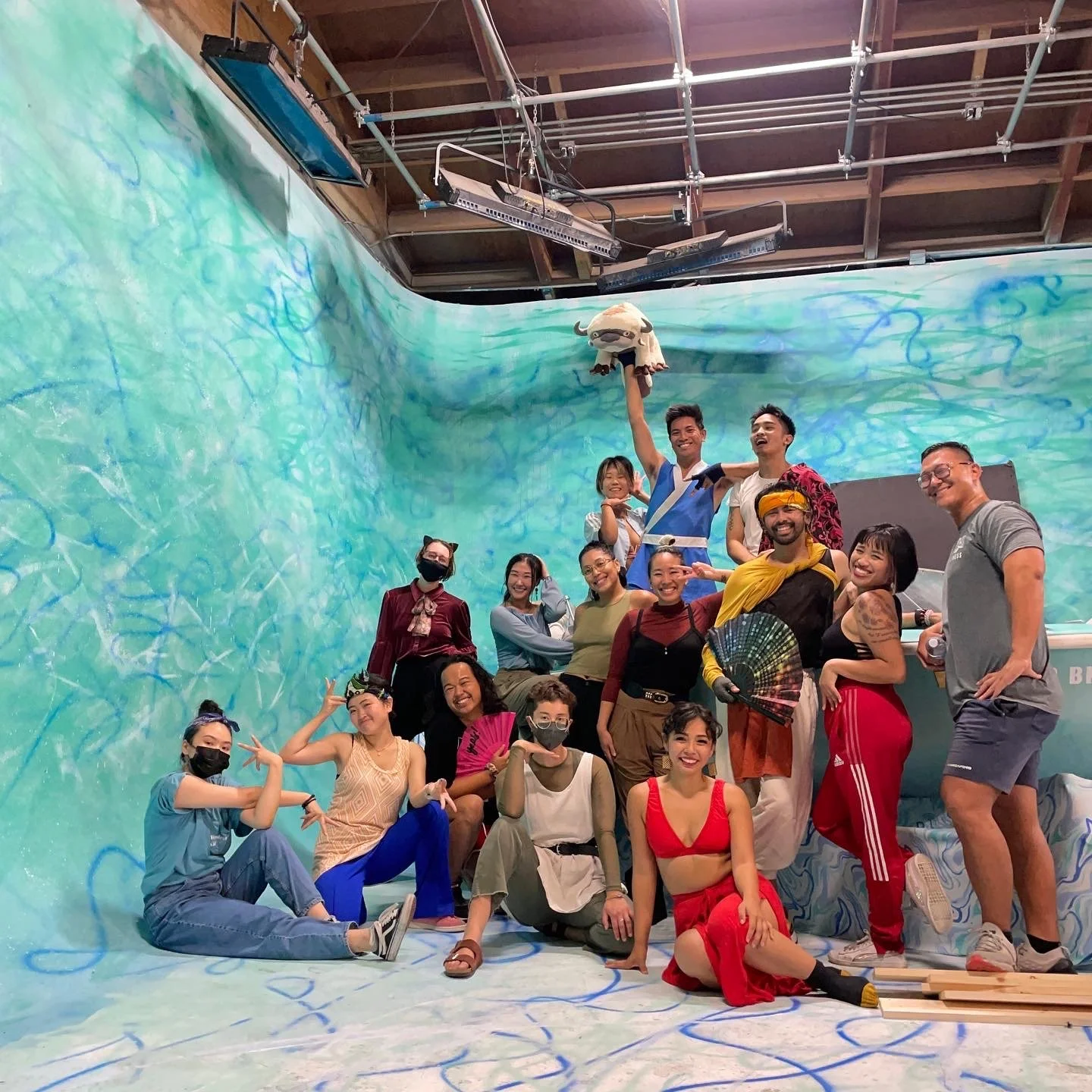 Group of people posing on a set with a large, colorful, abstract blue and green background, some in casual attire, some with costumes, smiling and making playful gestures.