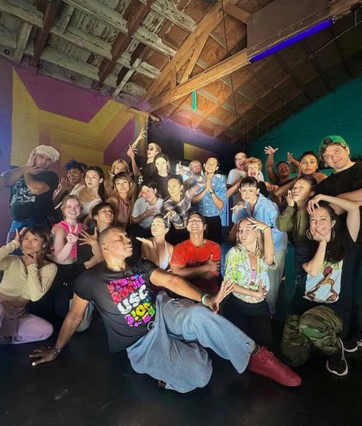 A group of people posing for a photo inside a colorful room with a high wooden ceiling, some people making funny gestures and expressions, and a large screen in the background.