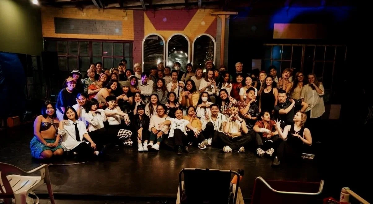 A large group of people posing for a photo on a stage in a dimly lit venue, with some individuals making hand gestures and smiling.