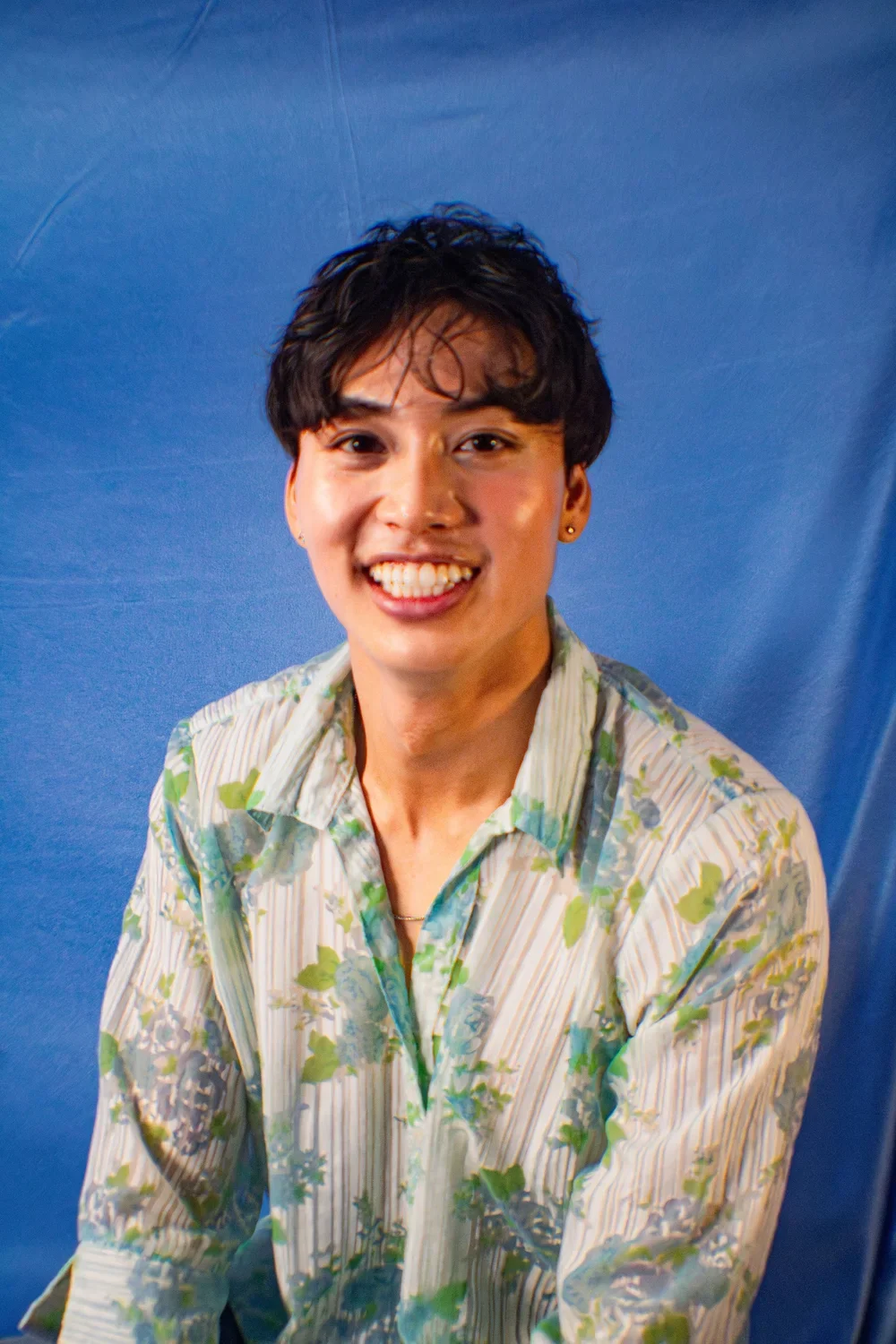 Young man smiling in front of a blue backdrop, wearing a floral patterned shirt.