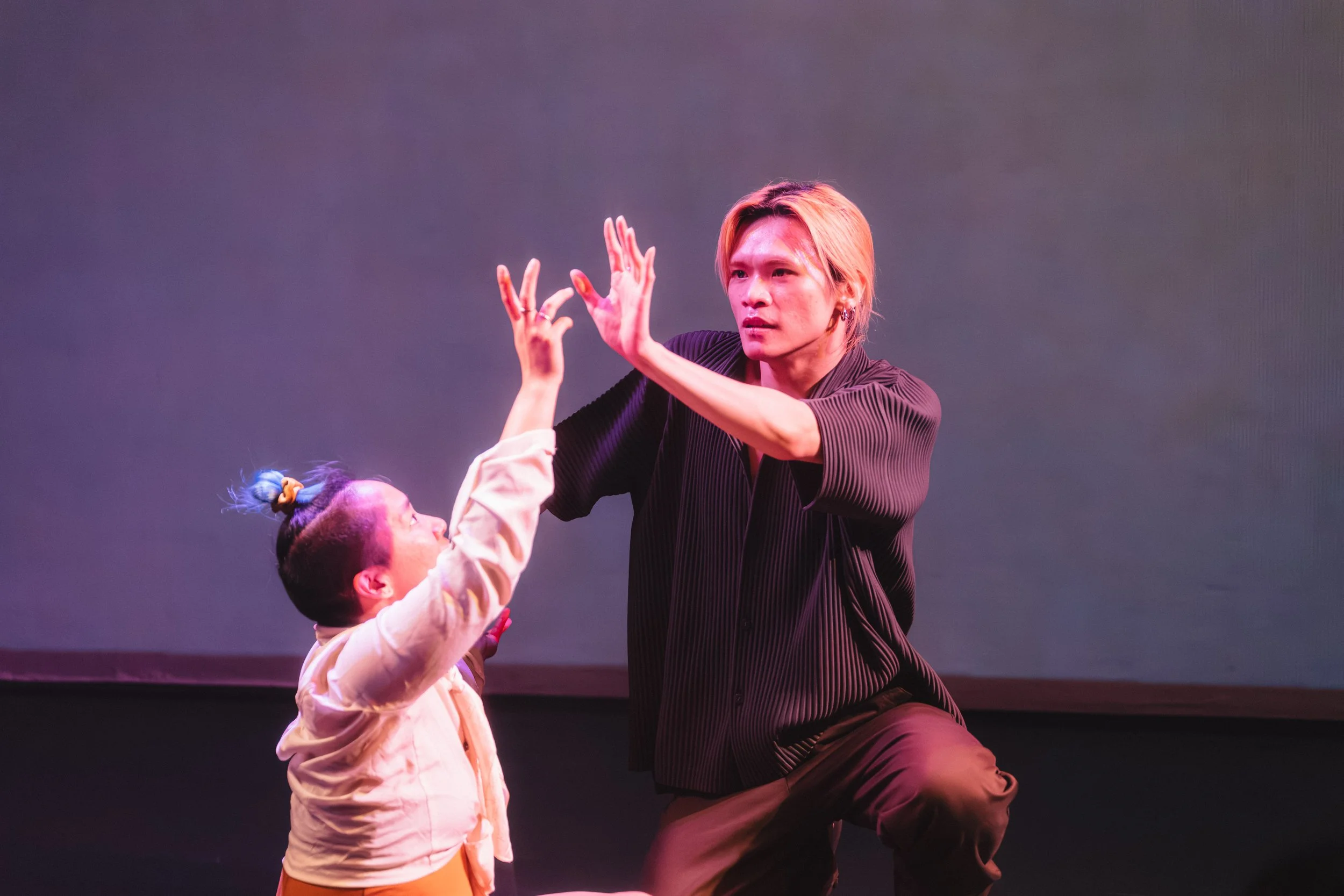 A performer with shoulder-length blonde hair, wearing a black striped shirt, is engaging with a child with colorful hair and a beige jacket, both on stage with a purple background. They are making expressive hand gestures, suggesting a lively performance or interaction.