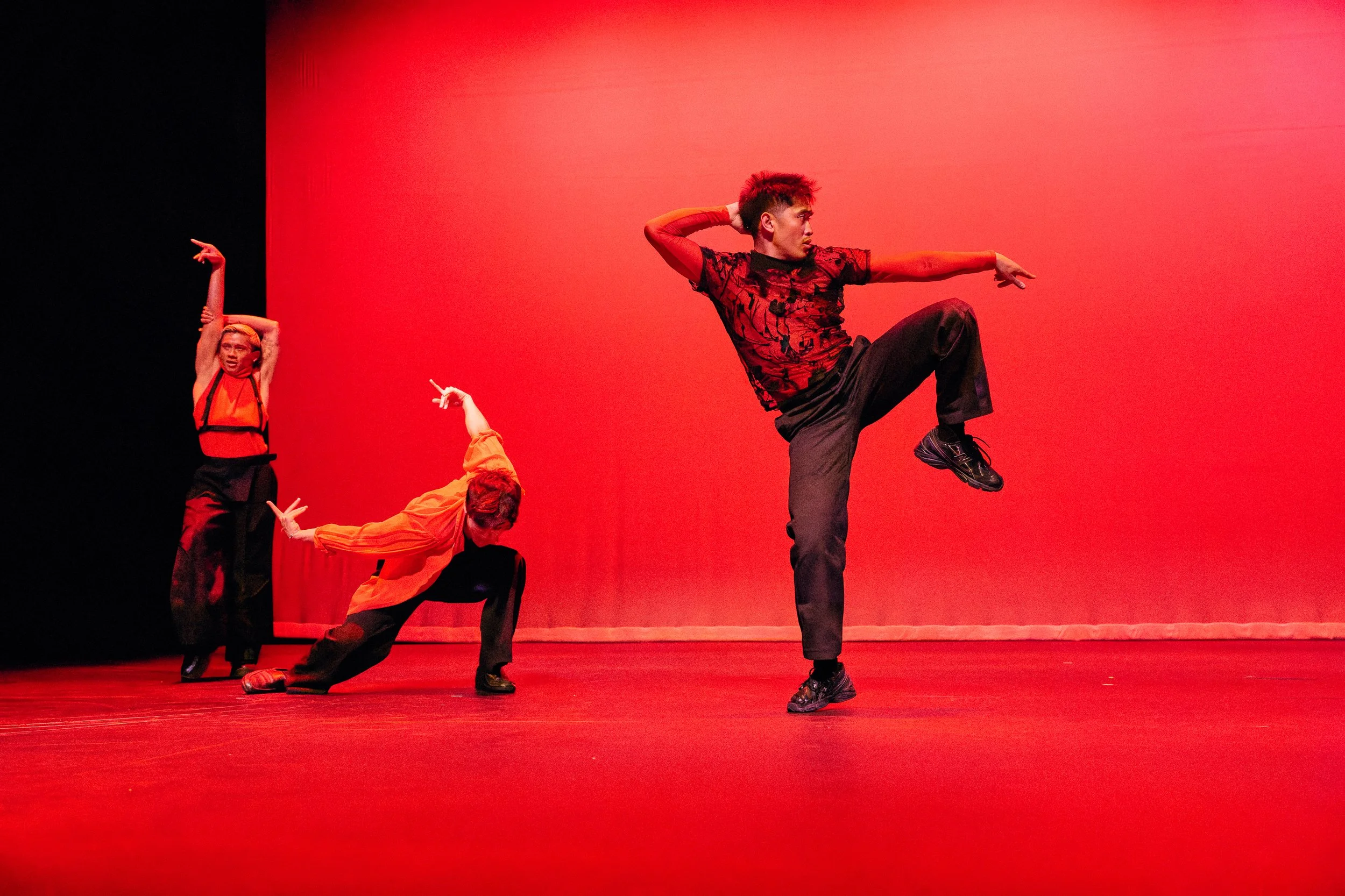 Dance performers on stage against a red background, with one standing on one leg and others in various poses.