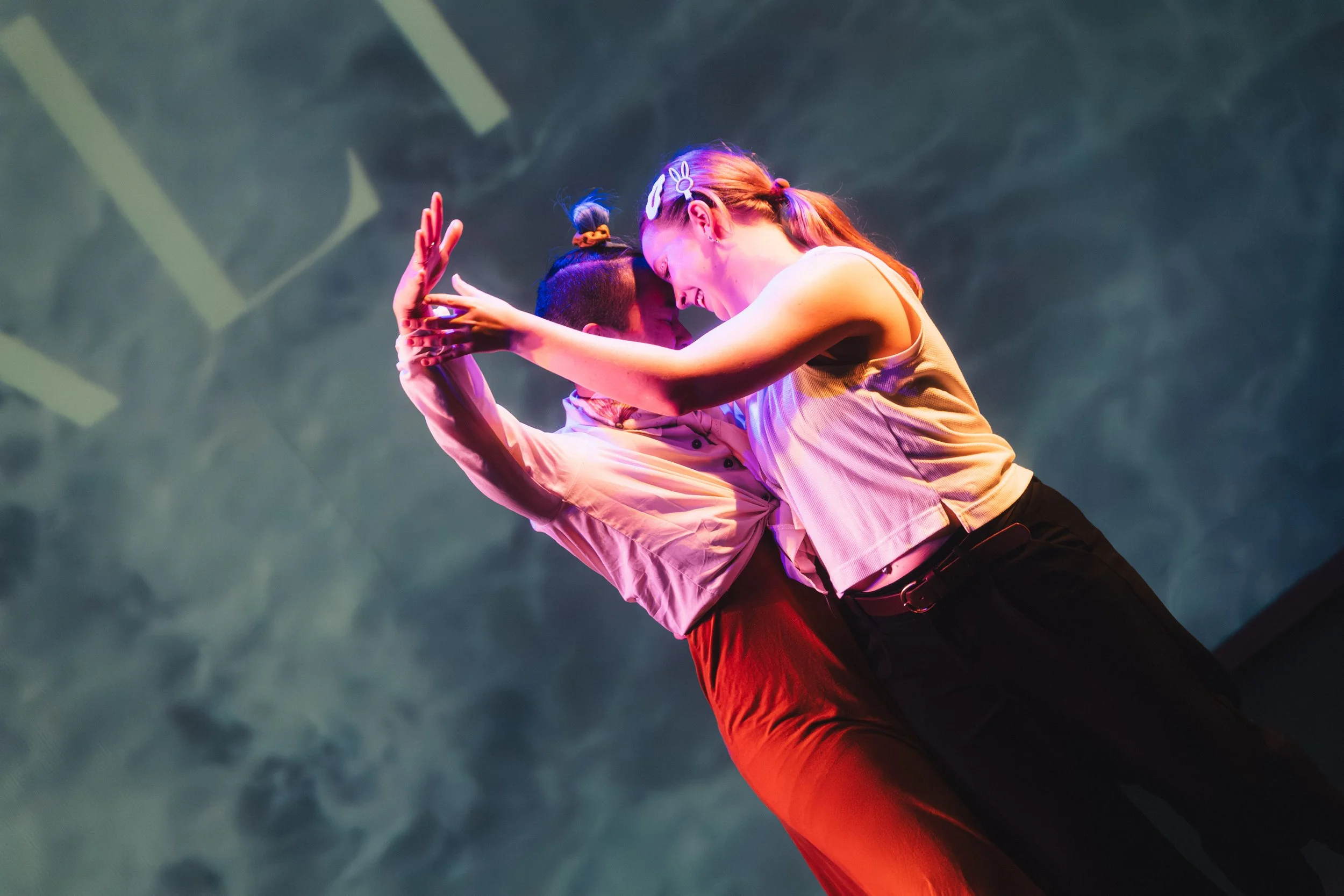 Two dancers perform a close partner dance on stage, with one dancer wearing a white shirt and the other in a yellow tank top, both smiling and leaning their foreheads together.